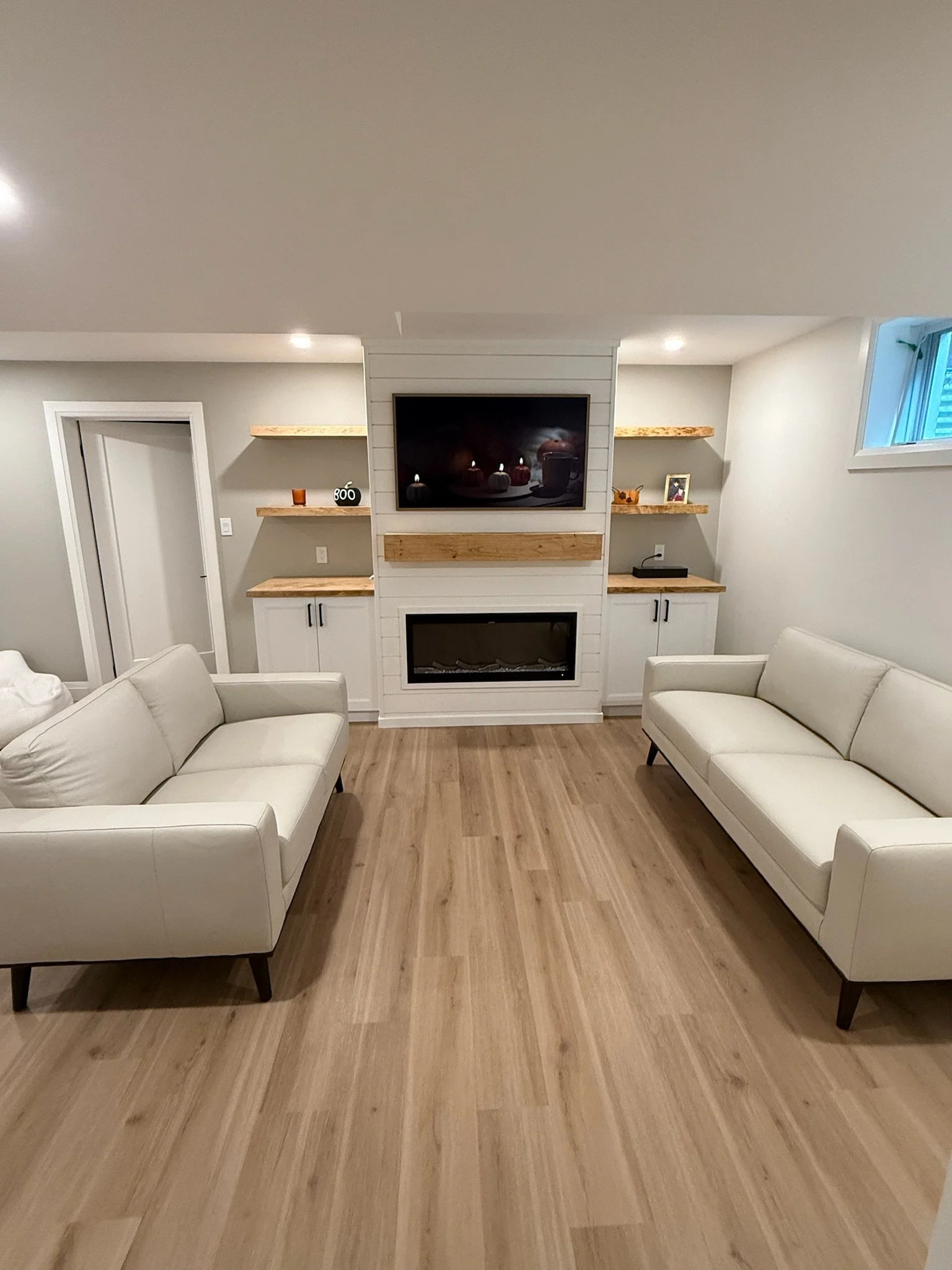 Living room with two white built in cabinets, a mounted flat-screen TV above a fireplace, and wooden shelves with decorative items against a light grey wall. London, ON.