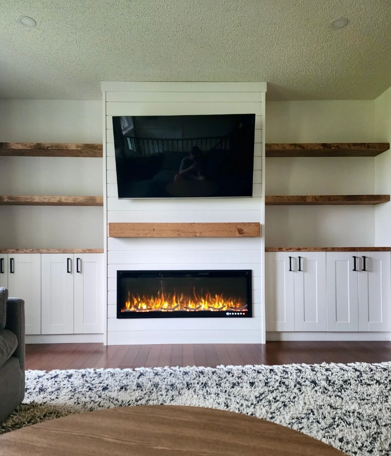 Living room with white built-in cabinetry, wooden shelves, a wall-mounted television, and an electric fireplace with flames visible beneath the TV.