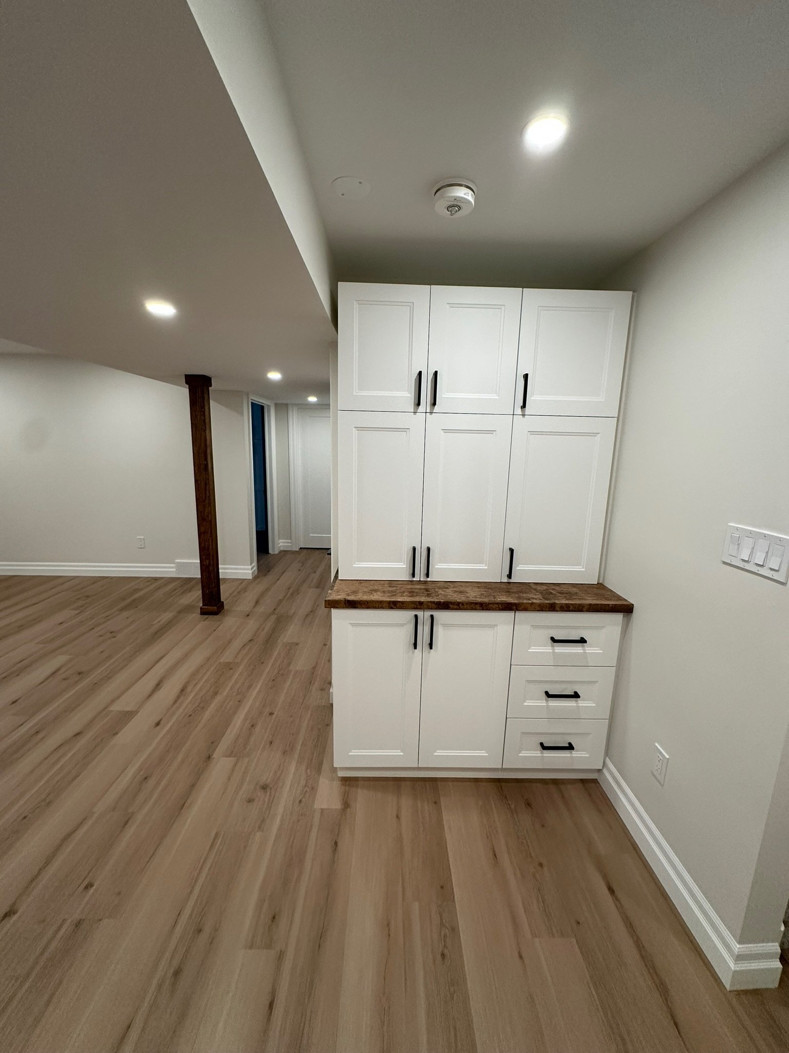 Interior of a room showing white kitchen cabinets, a wooden countertop, LVP flooring, a white wall with a light switch, and ceiling lights.
