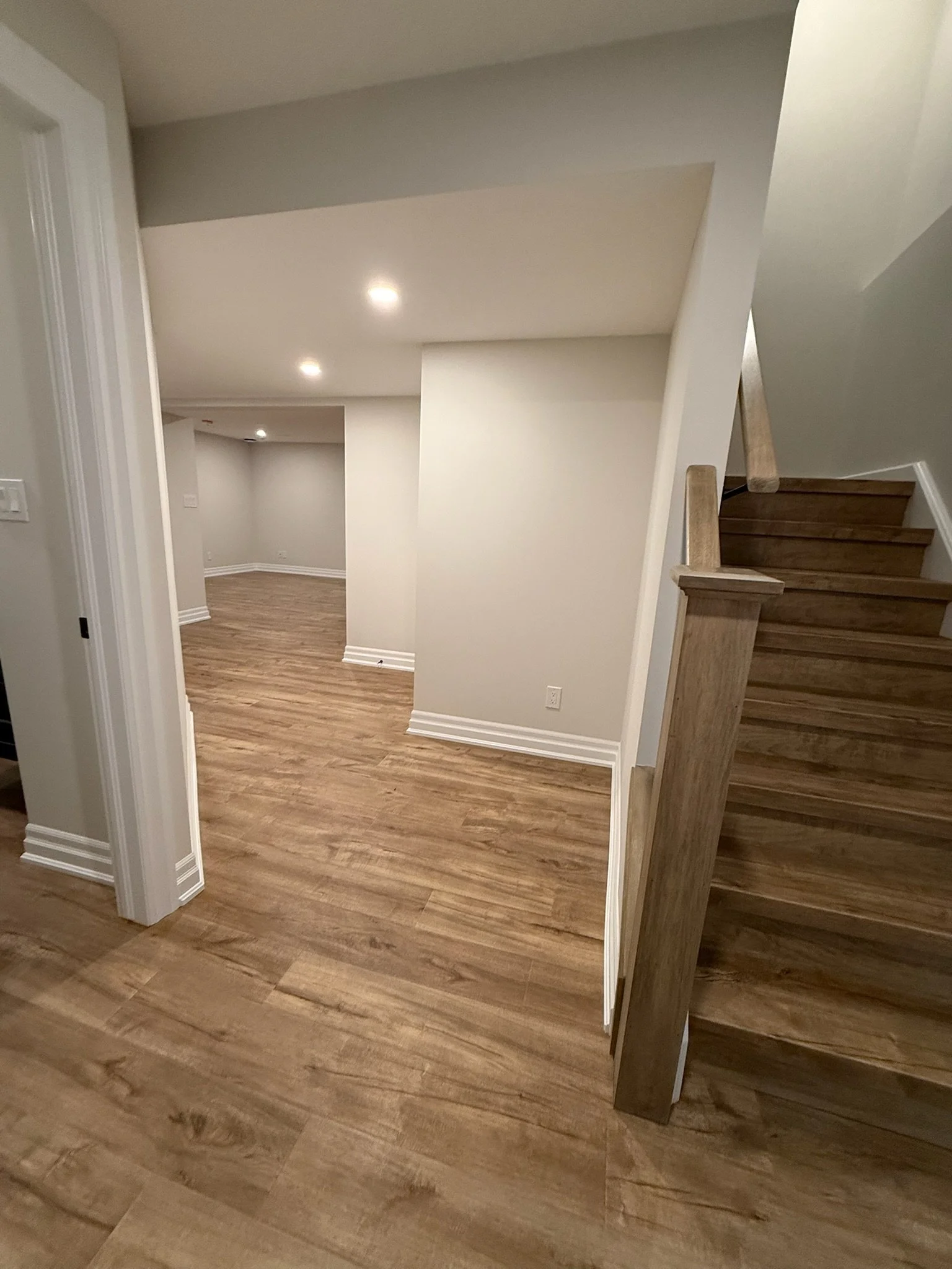 Interior view of a staircase with wooden steps and a wooden handrail, next to white walls and a partial view of a room with hardwood flooring.