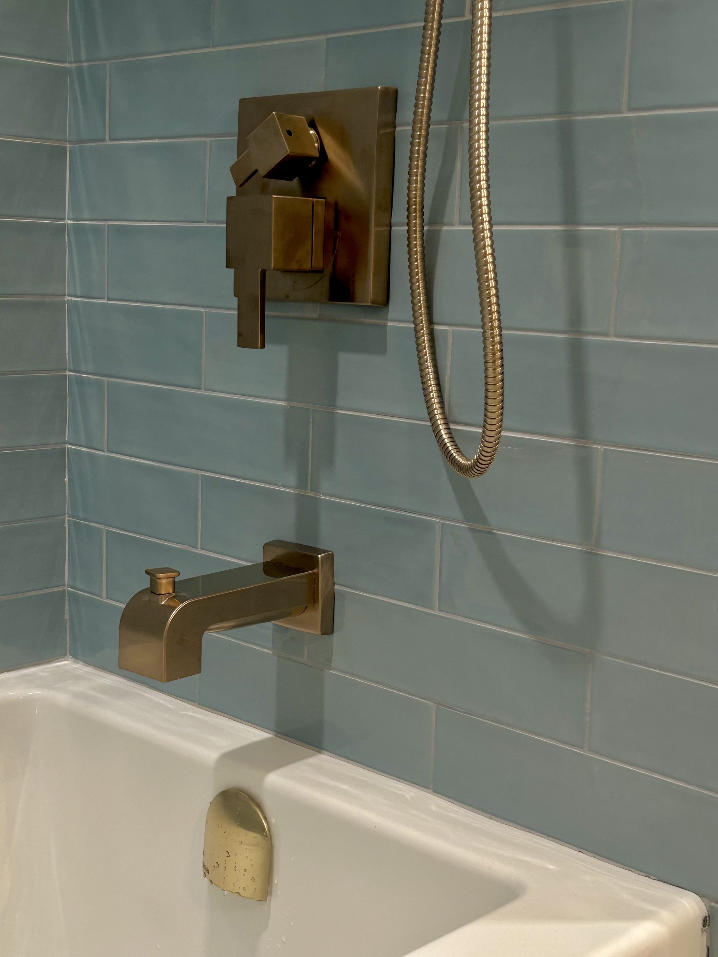 Close-up of a modern, gold-colored shower faucet with a matching handheld showerhead and hose mounted on a light blue tiled wall above a white bathtub.