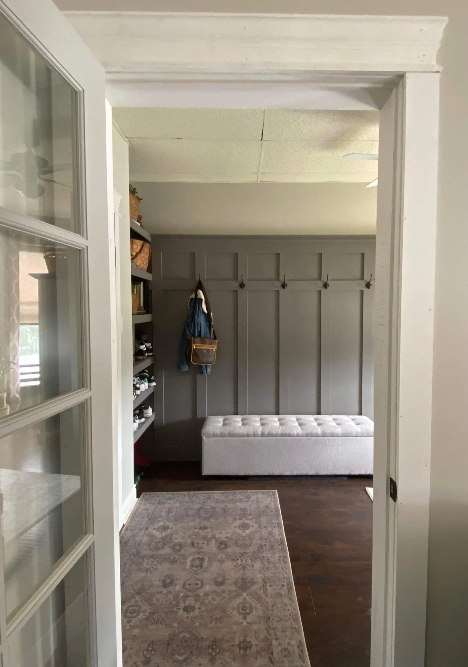 A hallway with a gray panel wall with hooks, a white tufted bench, a gray rug, and shelves with baskets on the left.