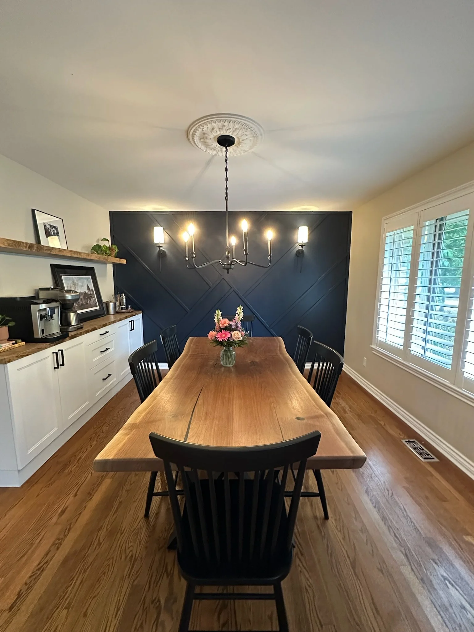 Dining room with a long wooden table, black chairs, a floral centerpiece, a black accent wall with wall sconces, a window with white shutters, and a ceiling light fixture.