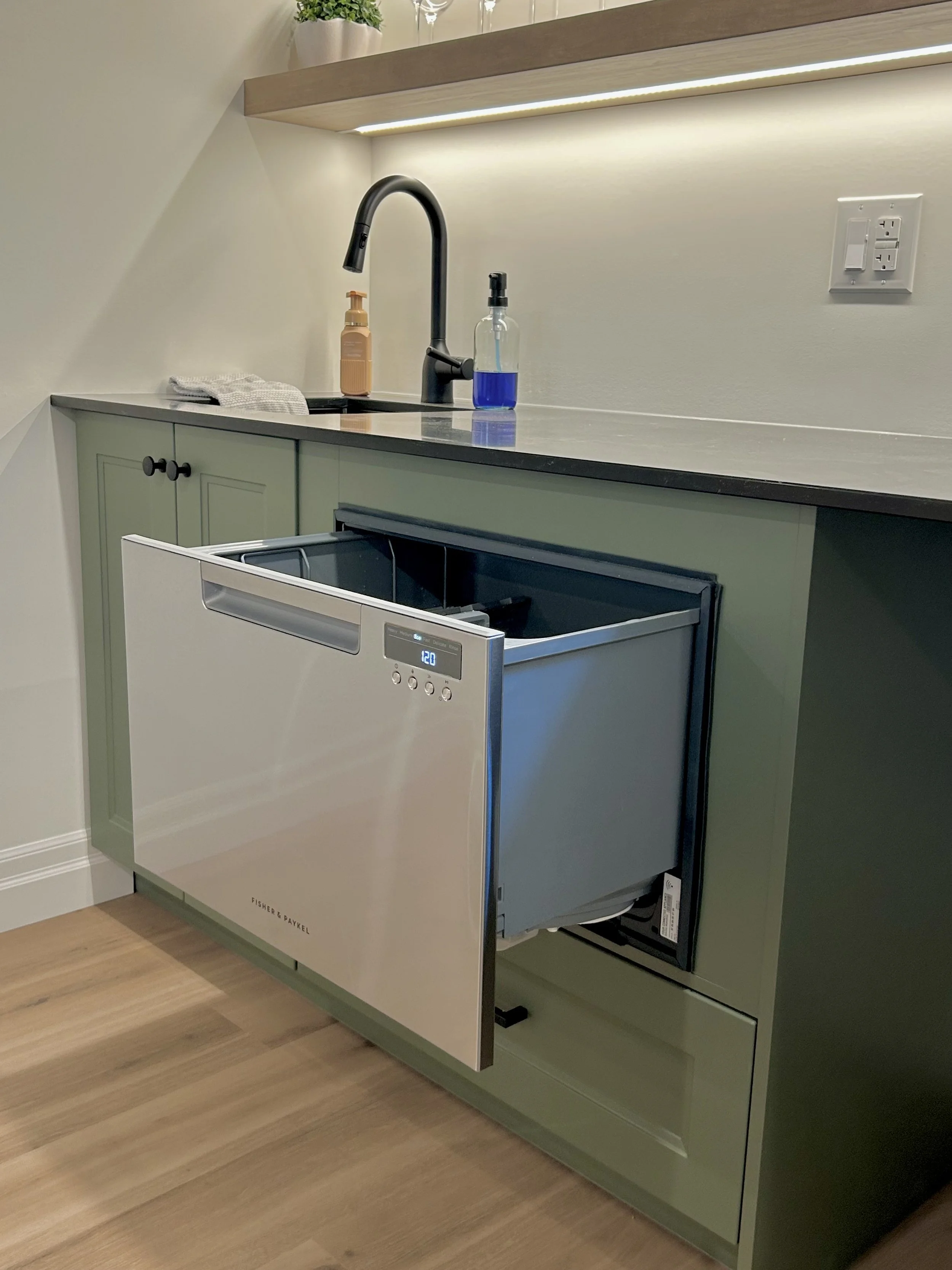 Kitchen counter with a black faucet, a soap dispenser, a blue liquid soap or sanitizer bottle, a dish towel, and a built-in dishwasher with an open door. There is a wooden shelf with a potted plant above the counter. A wall outlet and a light strip a