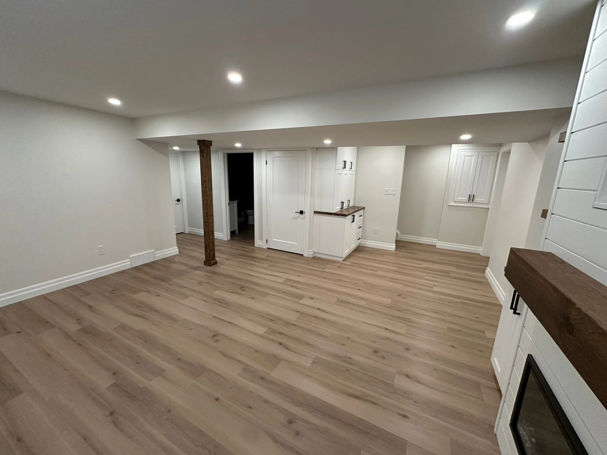 A newly finished basement with light-colored wood flooring, white walls, white cabinets, and recessed lighting in the ceiling. There is a wooden support post and a doorway leading to a bathroom with a toilet visible.