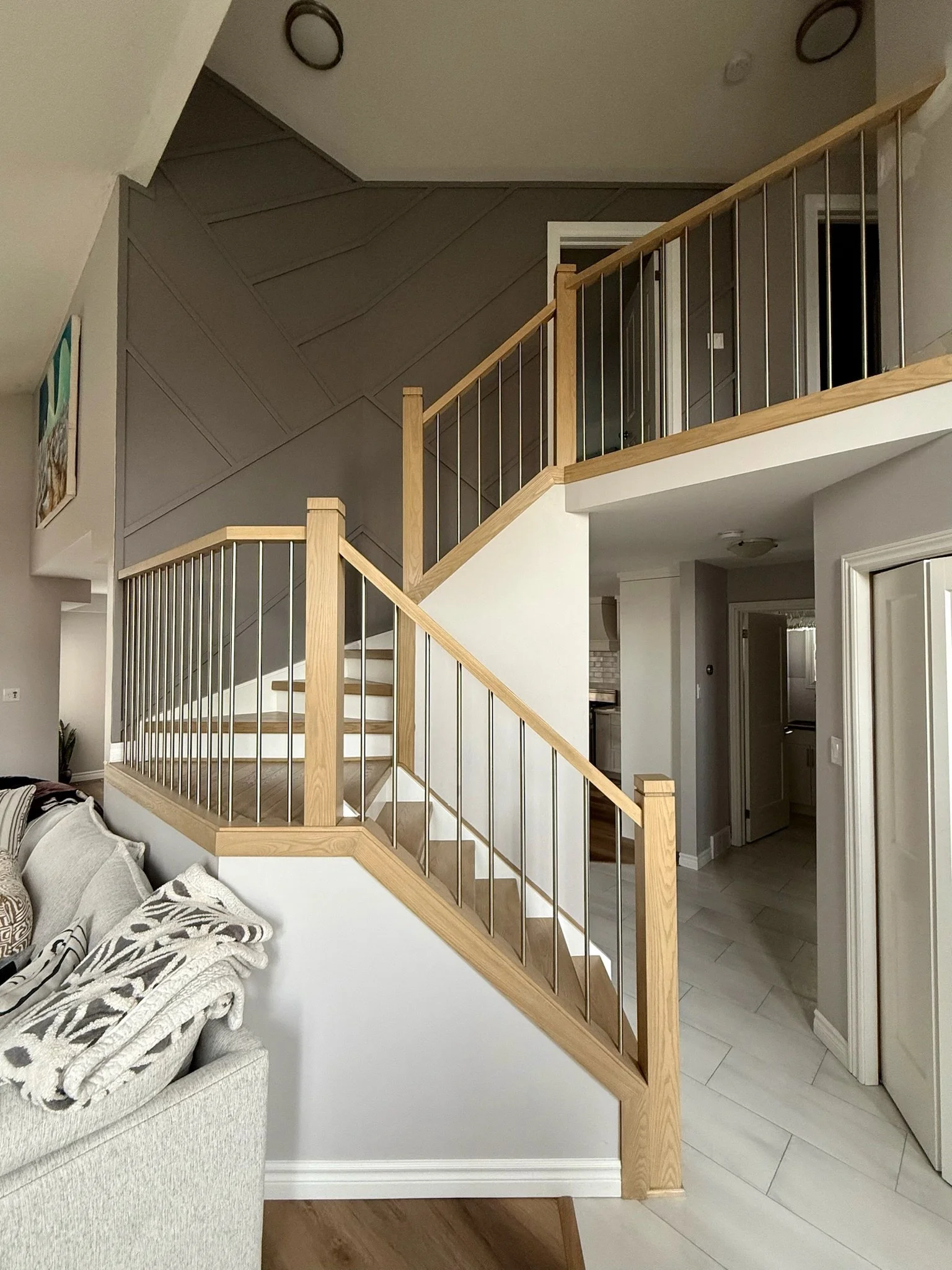 Interior view of a living space with a staircase. The staircase features light wood handrails and vertical metal balusters. Part of a light-colored sofa with patterned cushions is visible in the foreground. A gray wall with diagonal paneling is behin