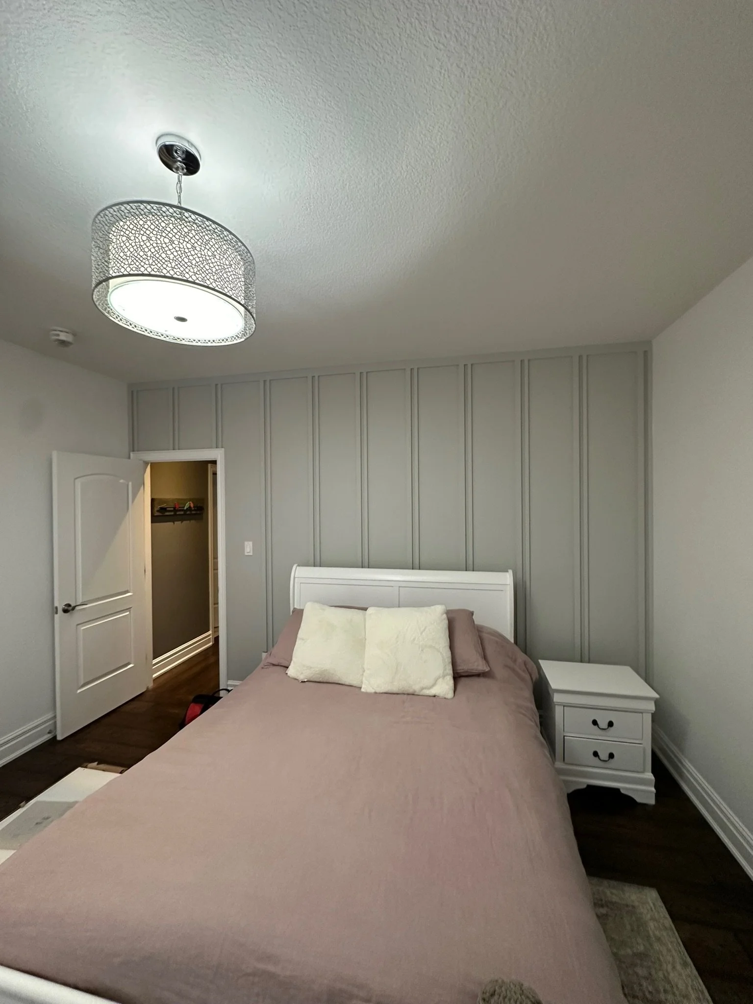 A bedroom with a bed covered in a pinkish-beige bedspread, white headboard, and two pillows, one white and one pale pink. There is a small white nightstand with two drawers beside the bed, and a gray wall with vertical paneling behind the bed. An ope