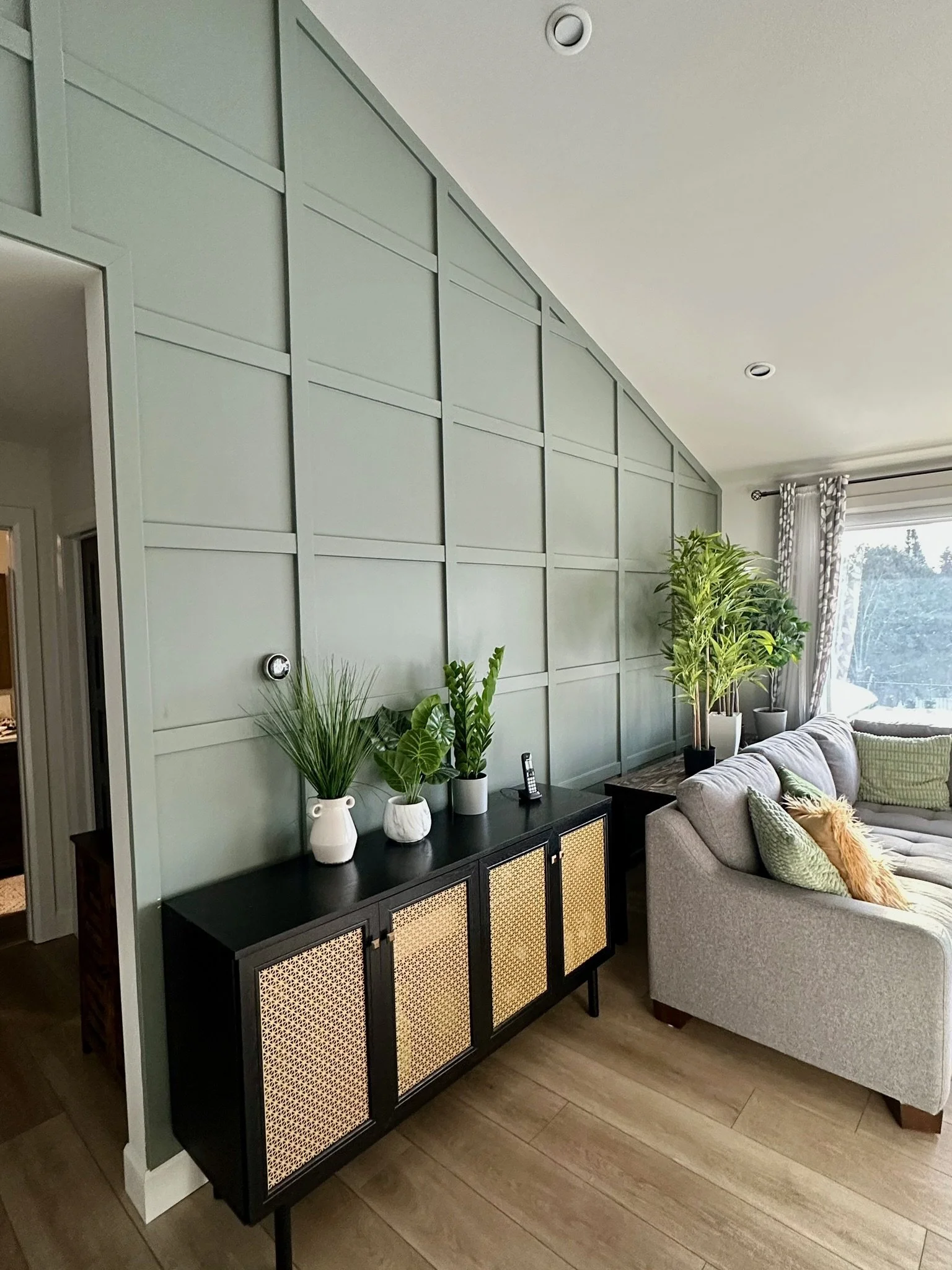 Living room with a sage green paneled wall, black sideboard with woven doors, and various potted plants. A beige sectional sofa with green, orange, and beige pillows, and a window with curtains in the background.