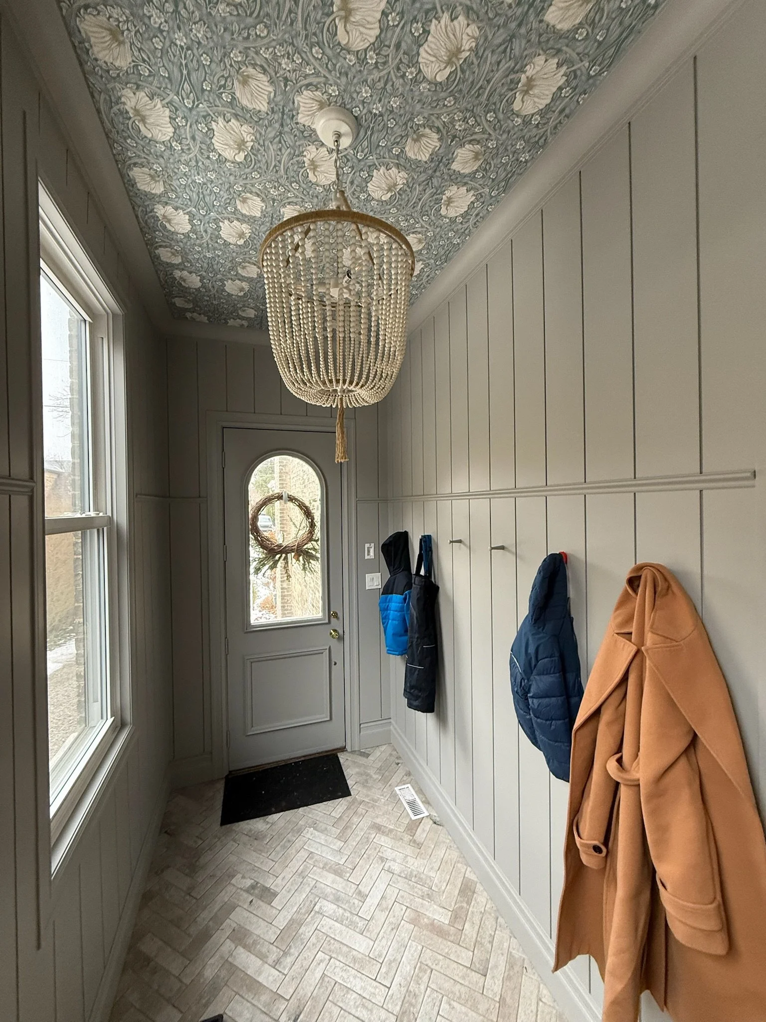 View of a hallway with a door and window, coat hooks, shiplap panels, on the wall with jackets hanging, and a decorative ceiling with floral wallpaper.