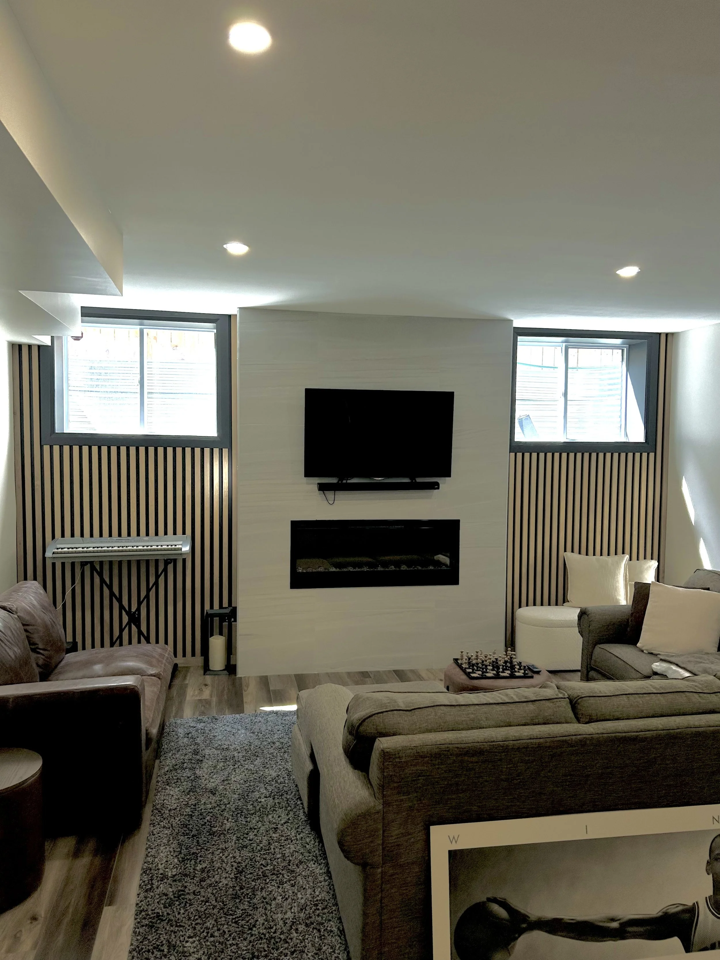 A living room with a television mounted on a wall above a fireplace, flanked by two windows with blinds, and beige sofas arranged around a small table with a chessboard. The room has wood flooring and a gray carpet.
