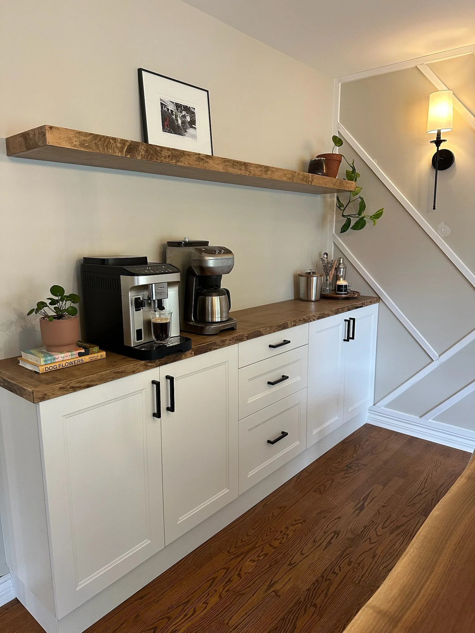 A coffee bar counter with white cabinets, a wooden countertop, wood floating shelf, a small plant, and books, situated underneath a floating shelf with a framed photograph and a potted plant, with a staircase and wall-mounted lamp in the background.