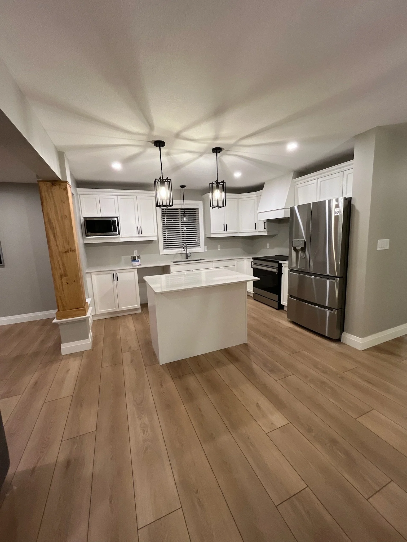 Modern kitchen with painted white cabinets, stainless steel appliances, a kitchen island, and wood flooring, illuminated by pendant lights.wood beam wrapped.