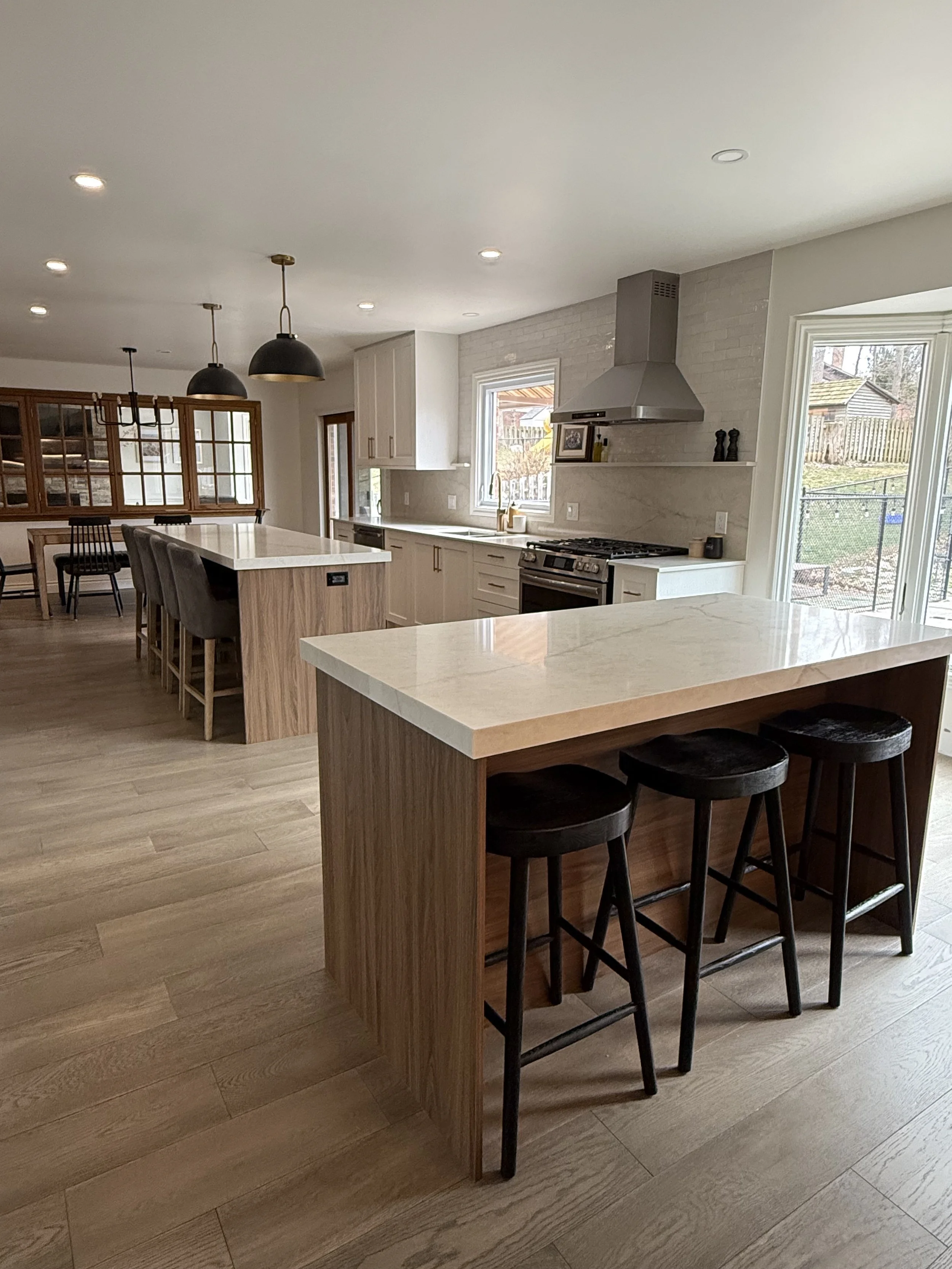 Modern kitchen with white cabinets, quartz countertops, stainless steel appliances, a large window, and a kitchen island with barstools. Light wood flooring. Adjacent dining area with a black chandelier and chairs. in London Ontario 