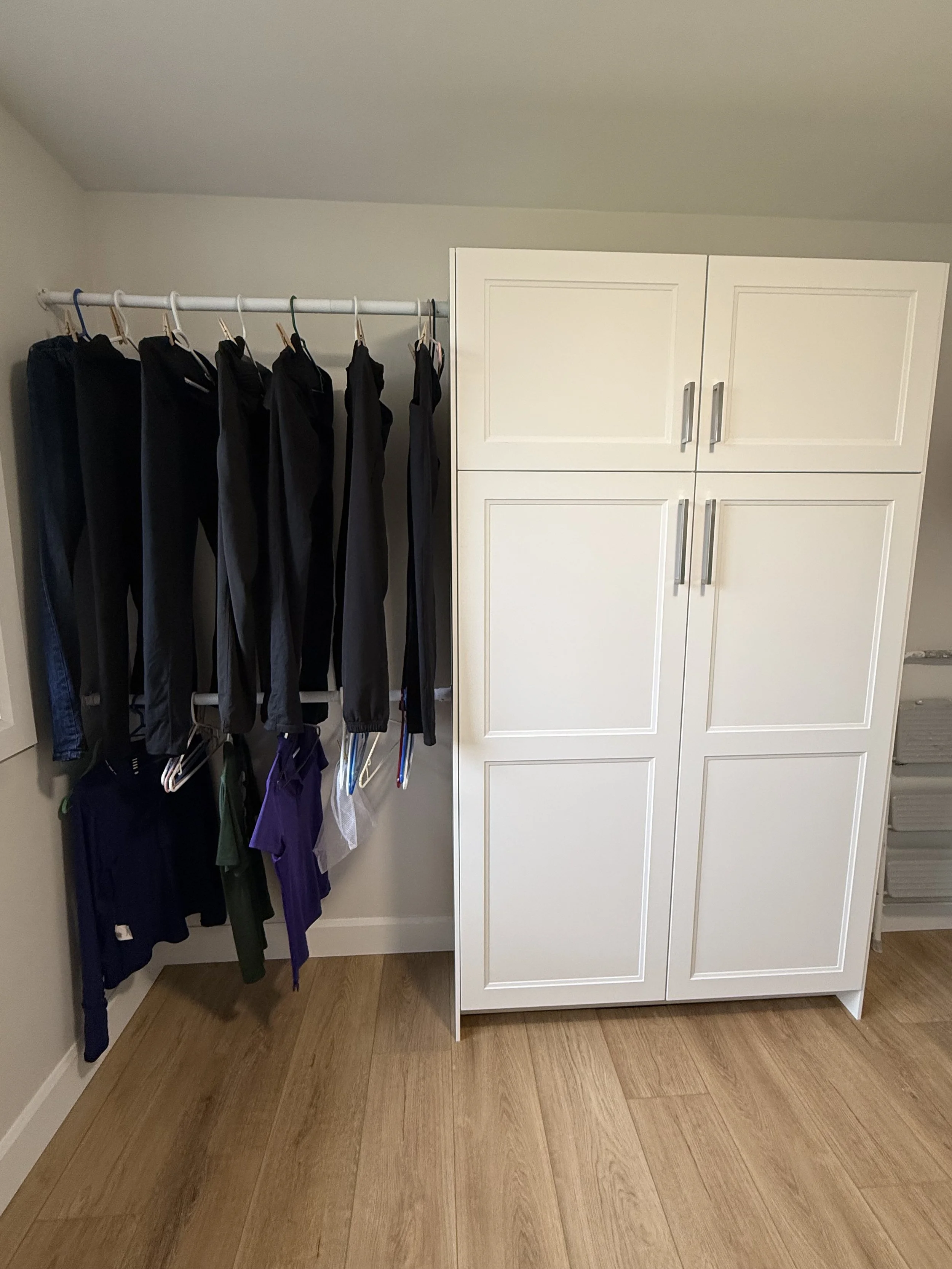 A white wardrobe with four doors and silver handles standing against a beige wall. To the left, there is a white clothing rack holding black pants and some shirts. The floor is hardwood.