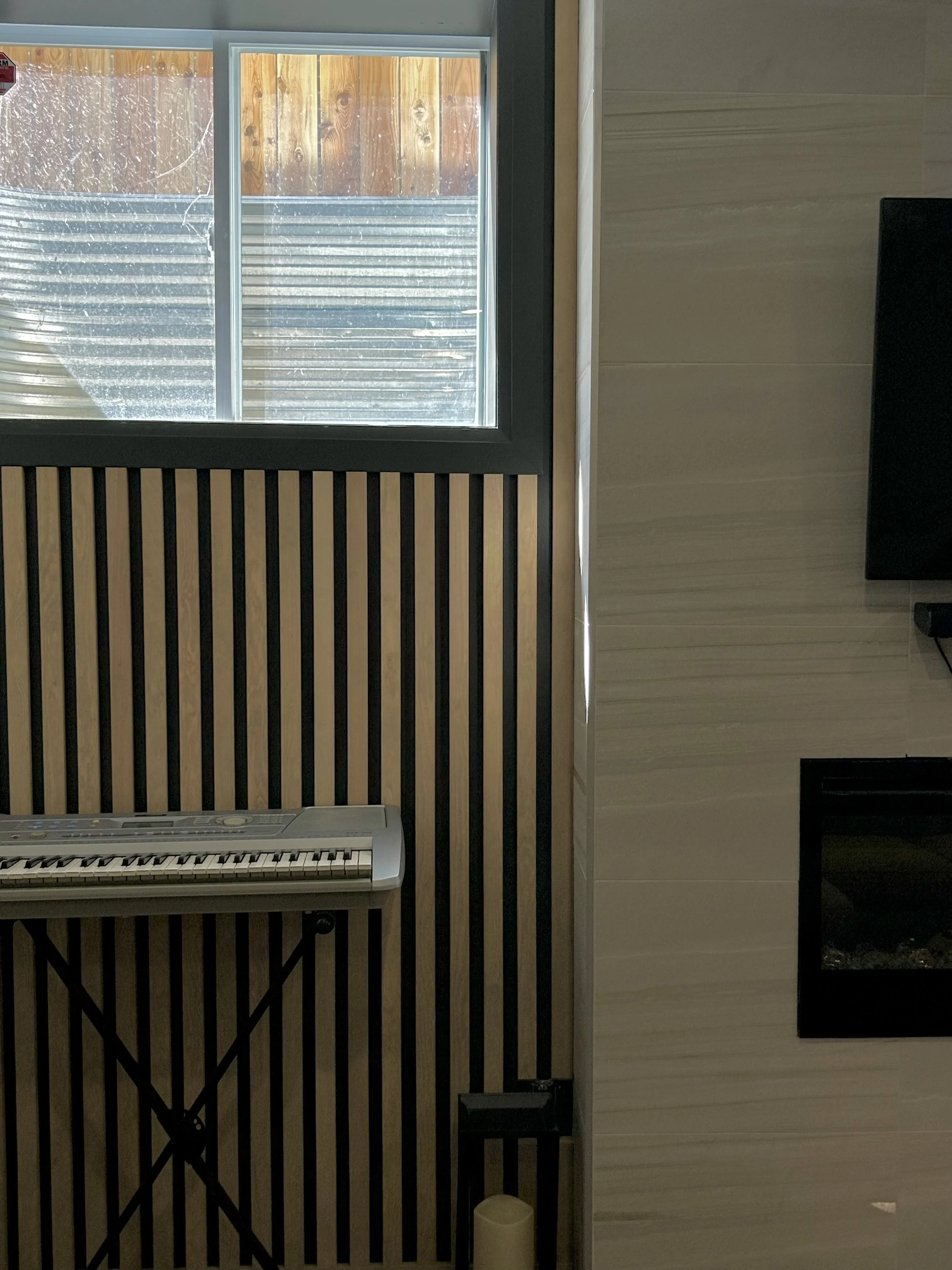 A section of an interior room showing a window with a wooden fence behind it, a black and beige slatted wooden panel, a white electronic keyboard, and beige horizontal wall panels with a black electrical device on the right side.