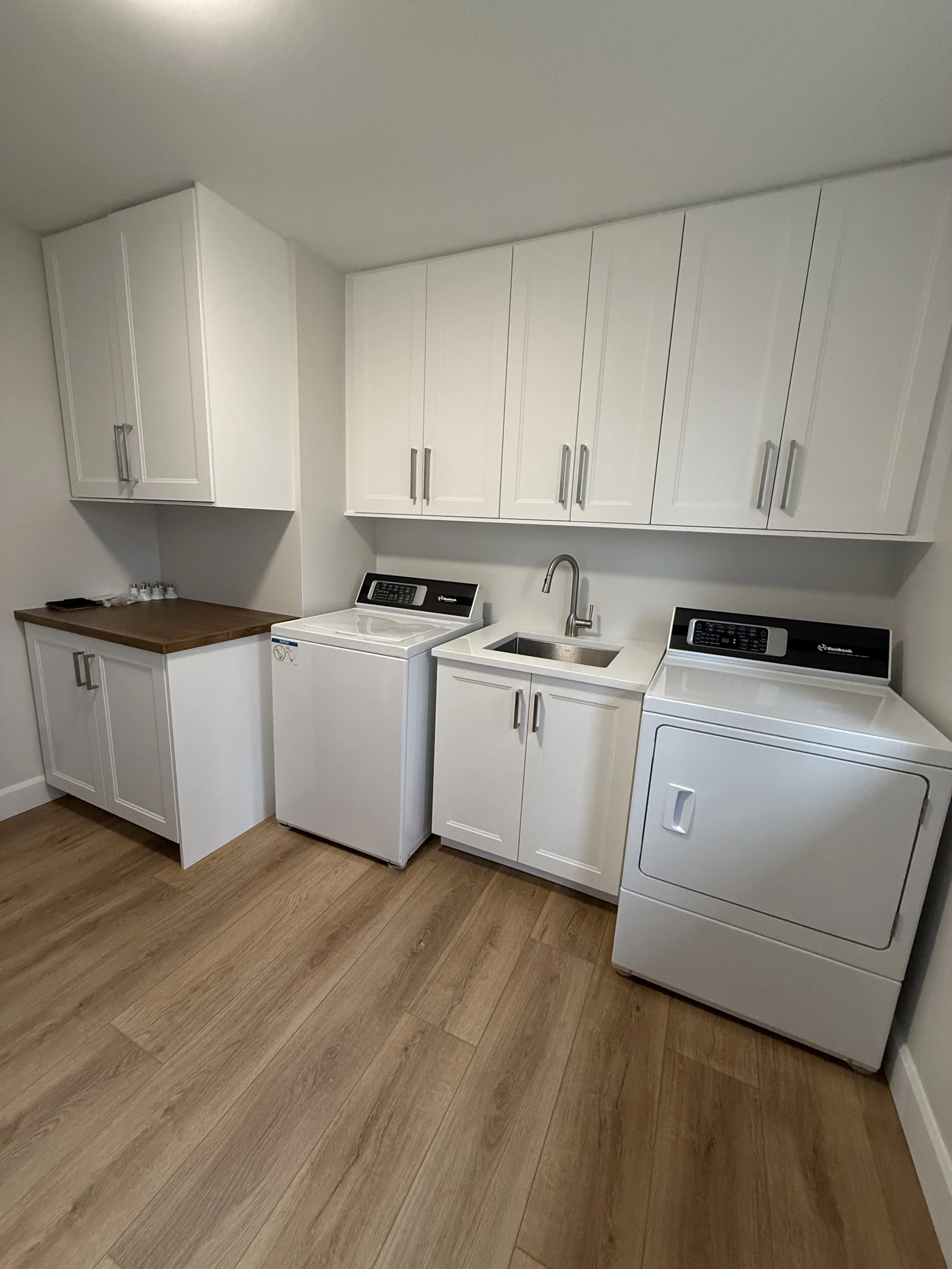 A laundry room with white cabinets, a washing machine, a dryer, and a utility sink with quartz counter and a faucet. There is a wooden countertop and a set of small bottles on the countertop. The room has wood flooring.