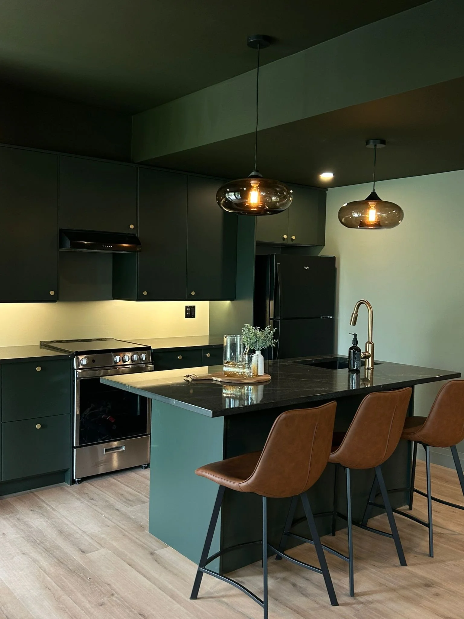 Modern kitchen with dark green cabinets, black countertops, stainless steel appliances, pendant lighting, and brown chairs around a kitchen island.