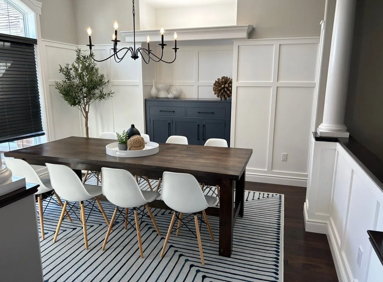 A modern dining room features wainscoting accent walls with a wooden table, white chairs, a black chandelier, a blue sideboard, and decorative objects on the walls and table, with a striped area rug and dark hardwood flooring.