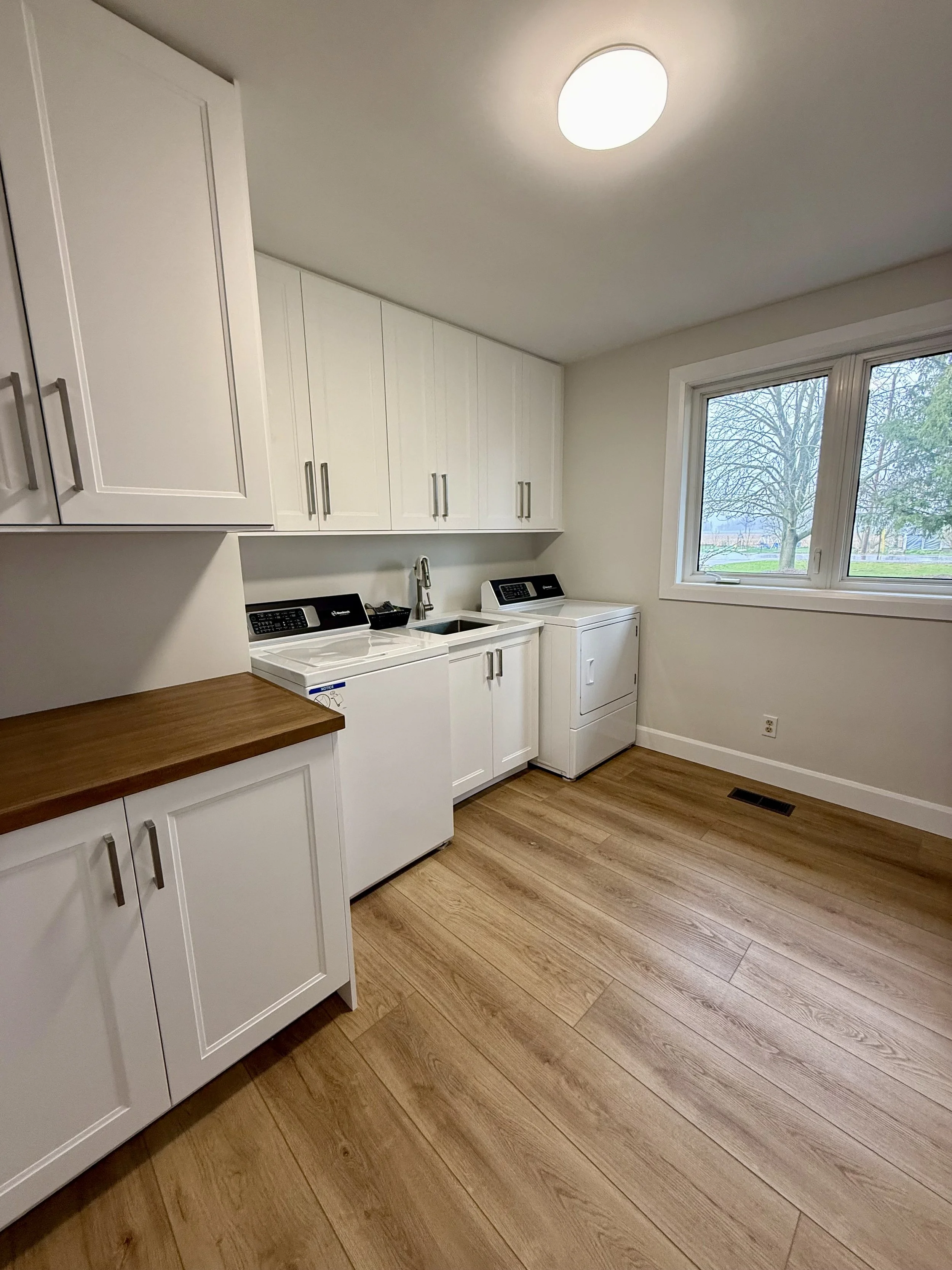 A laundry room with white cabinets, a washing machine, and a dryer by a window showing trees outside. The room has hardwood floors and a ceiling light.
