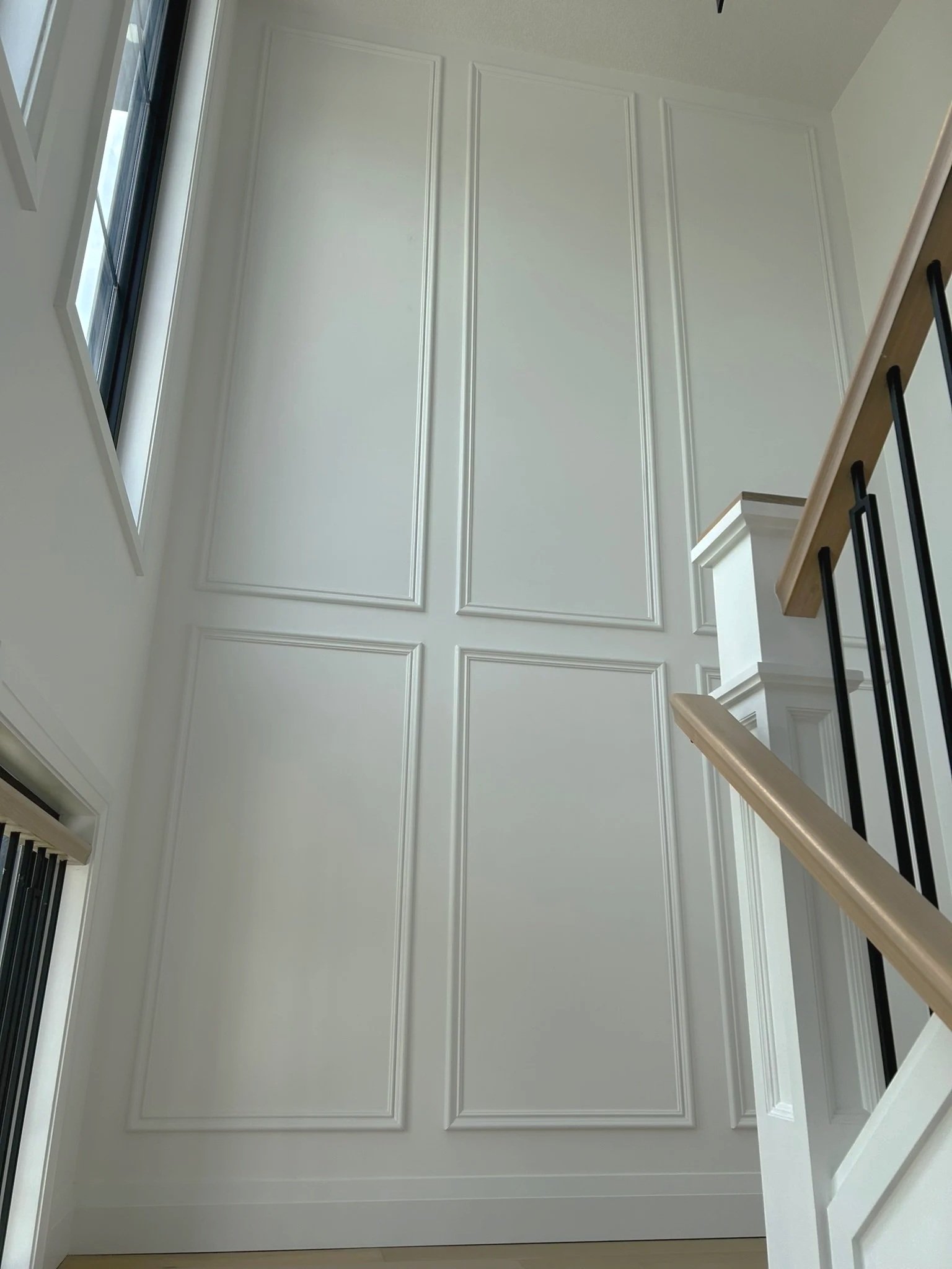Interior view of a staircase with a white wall featuring decorative molding, a wooden handrail, and black metal balusters, with a window on the left allowing natural light to enter.