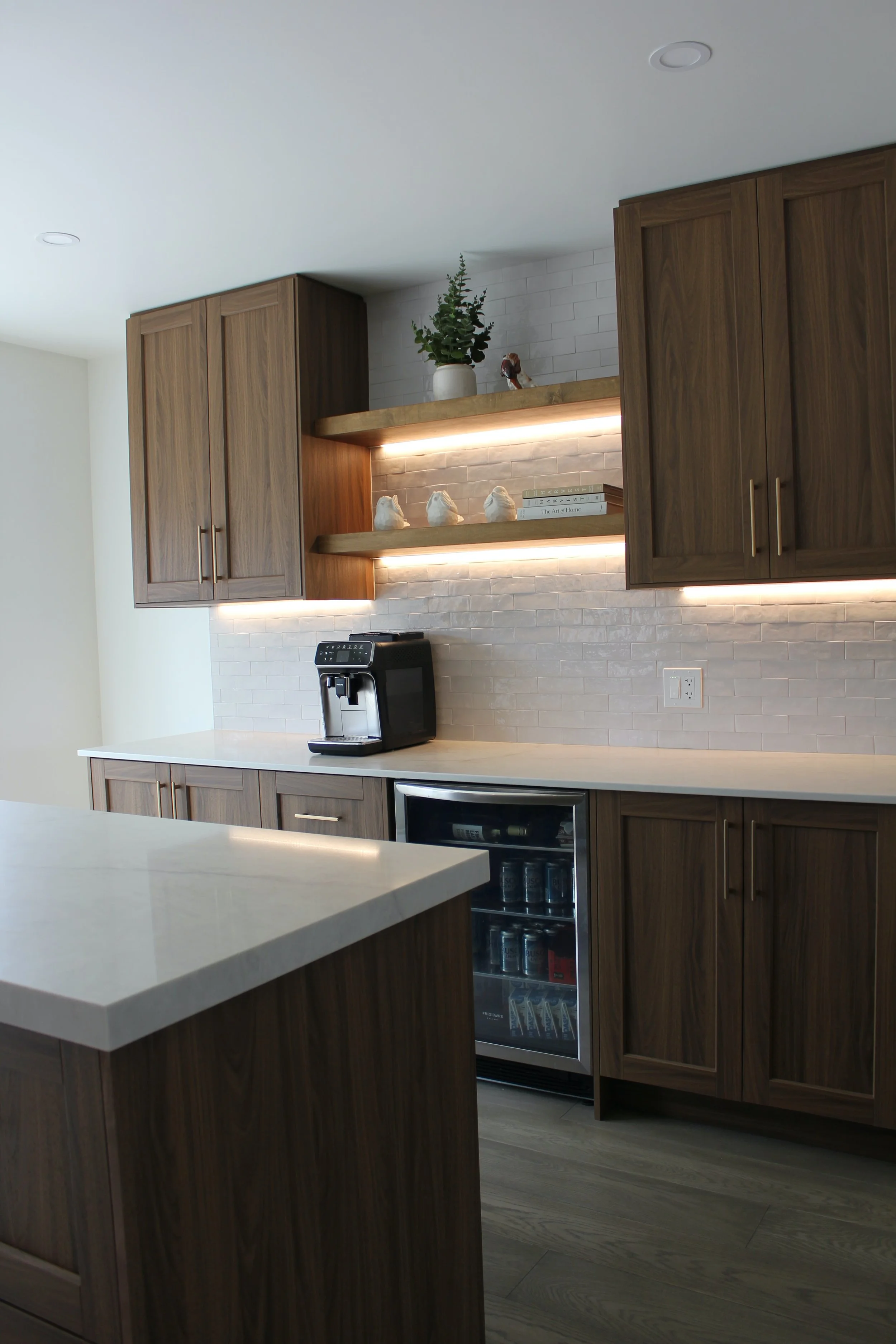 Modern kitchen with wooden cabinets, taj mahal countertop, coffee maker, small wine fridge, and open shelves with decorative items and books, illuminated by under-cabinet lighting.