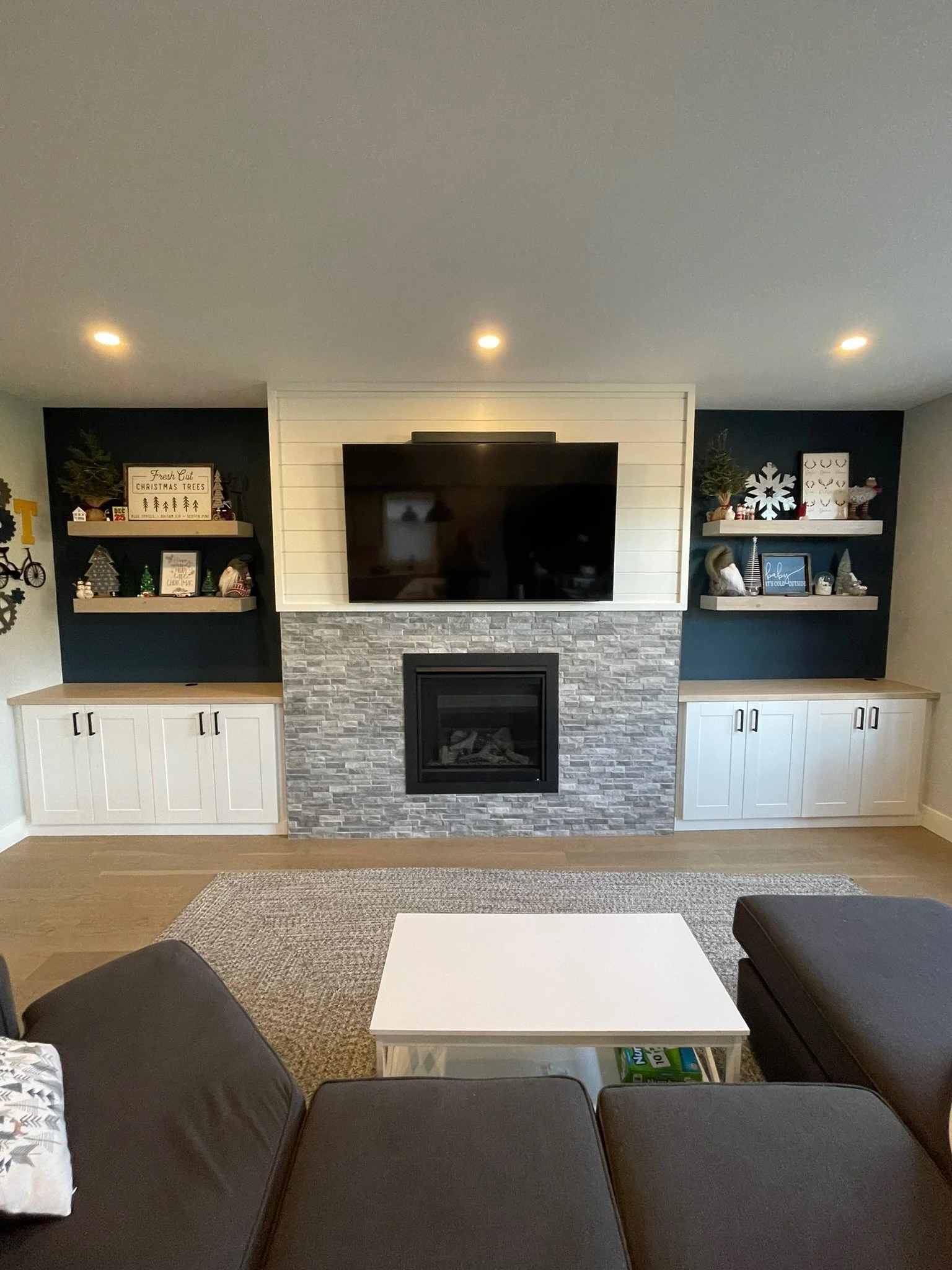 Living room with a stone fireplace, mounted TV, white and navy blue shelves decorated with holiday and Christmas decorations, cushions on a couch, a white table, and a beige rug.