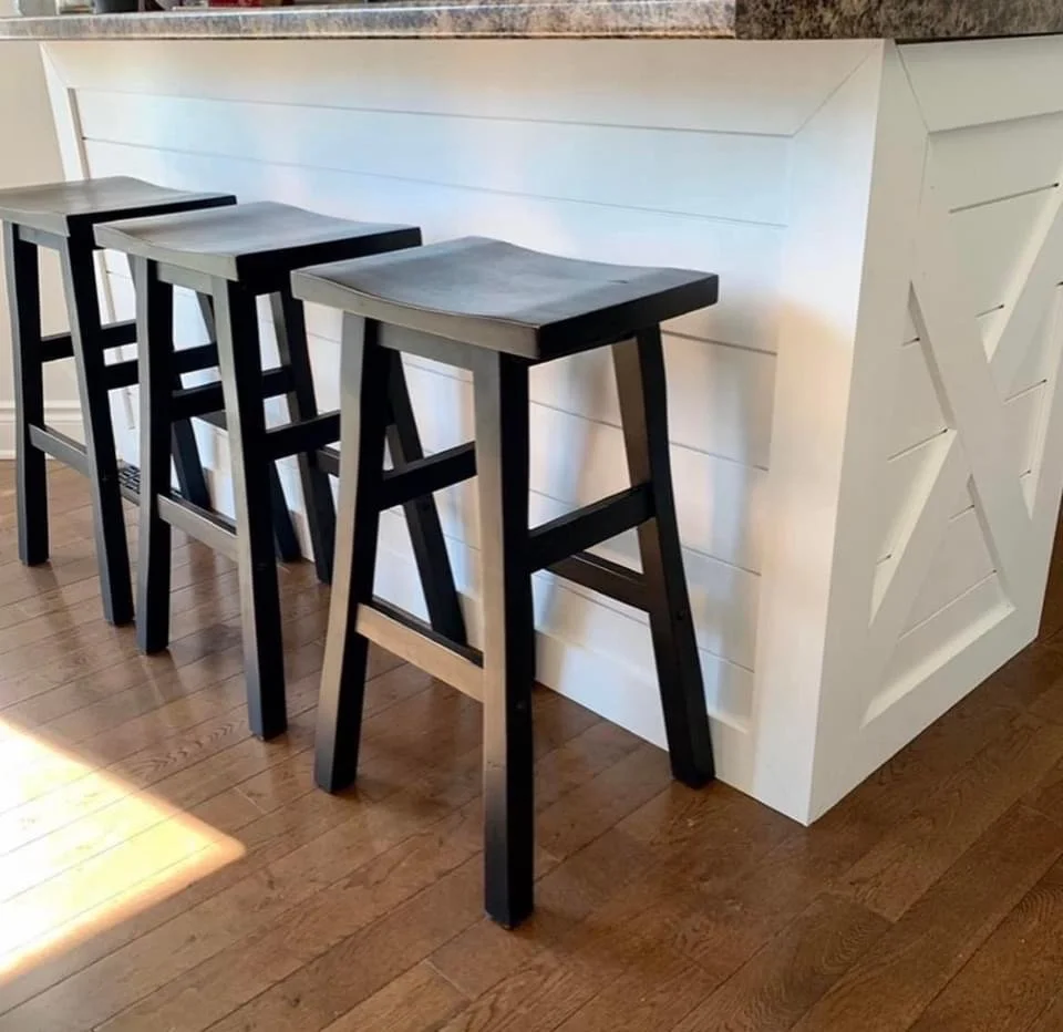 Three black and gray bar stools lined up against a with white wood paneling on the back of the island and X design on the side. placed on a wooden floor.