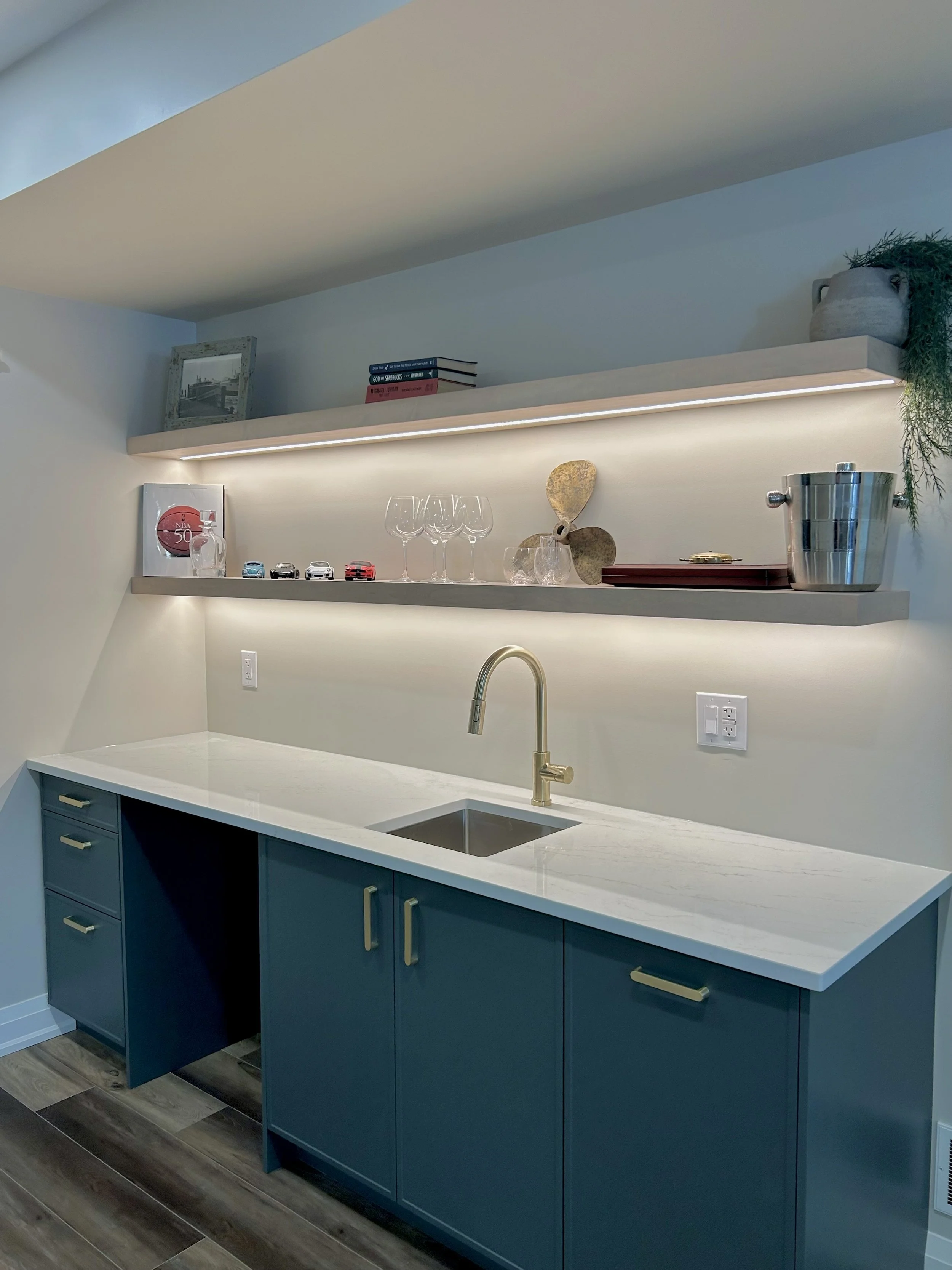 A modern kitchen with navy blue cabinets, a white countertop, a stainless steel sink, and open shelves displaying wine glasses, decorative items, and small model cars. The shelves have built-in lighting, and the floor is LVP.