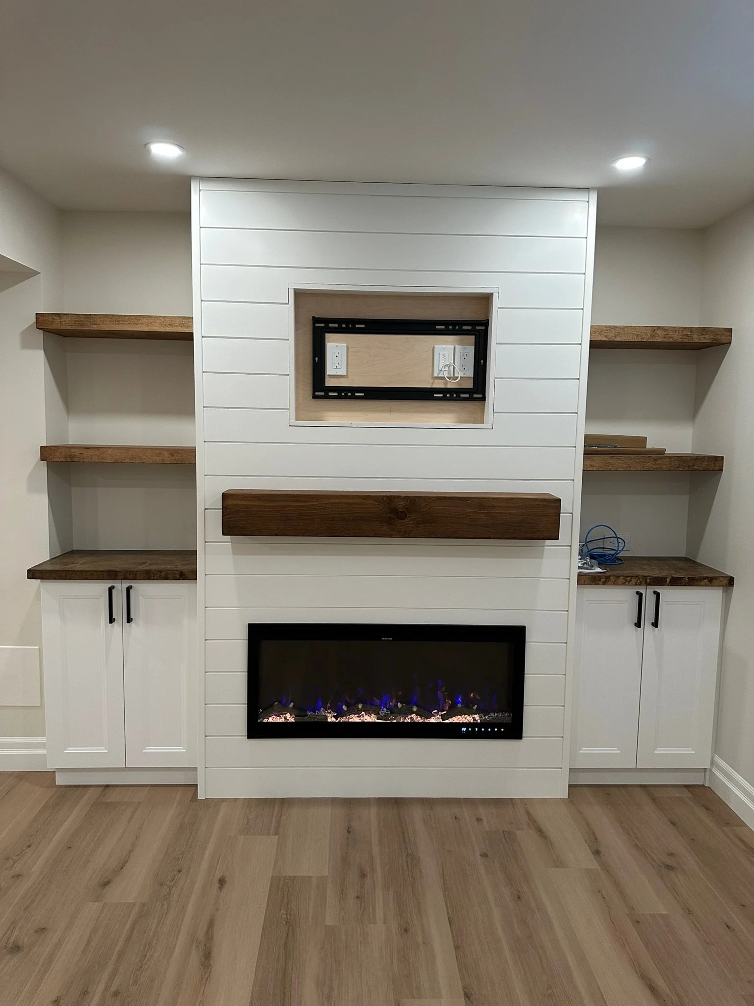 Empty built-in entertainment center with a modern electric fireplace at the bottom, white shiplap walls, wooden shelves, and hardwood floors.