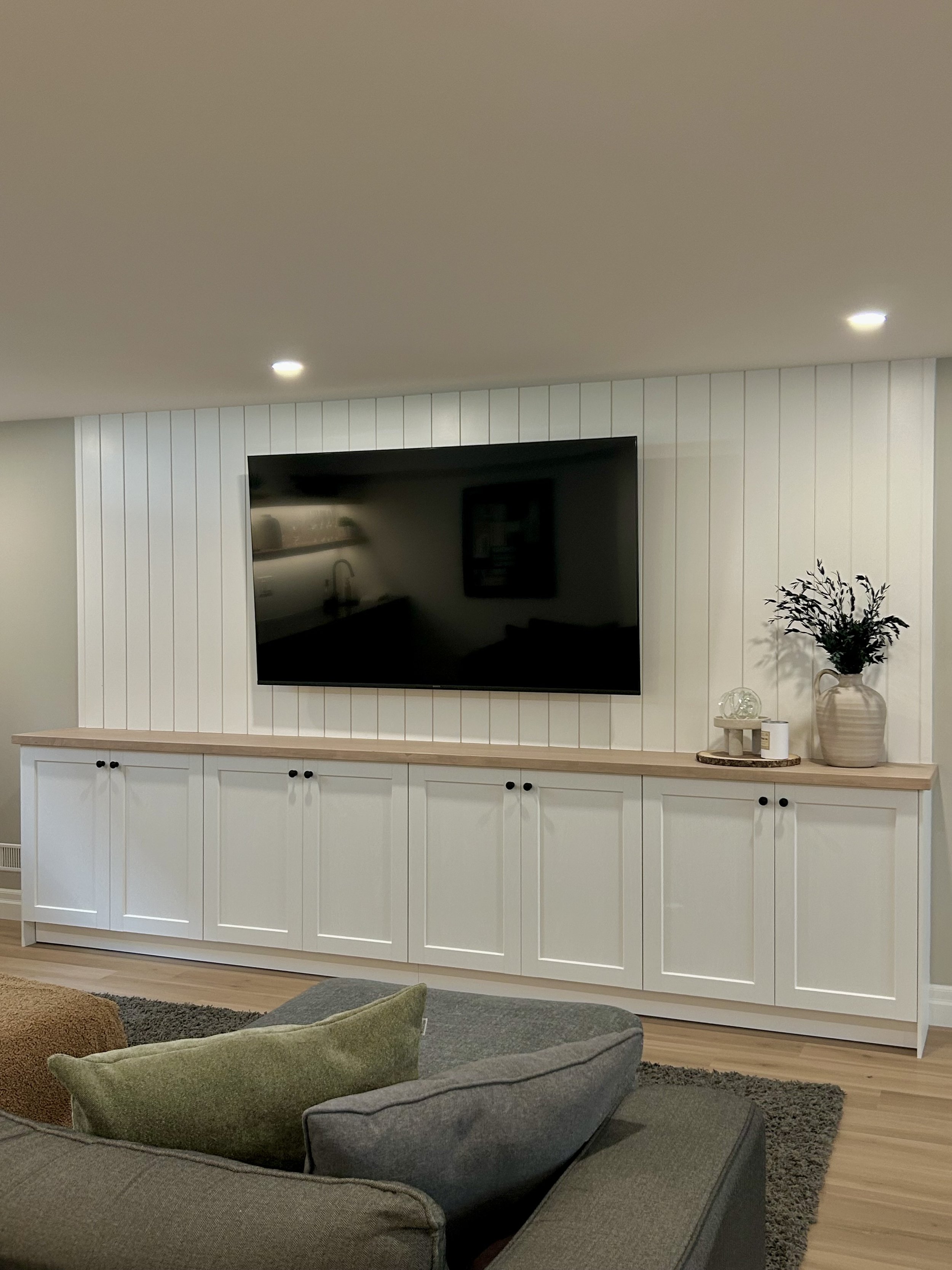 Living room area with a wall mounted flat-screen TV, white cabinet with black knobs, and decorative items on top, including a vase with artificial plants, a small sculpture, and a framed picture. A gray sofa with green and brown pillows, a black rug,