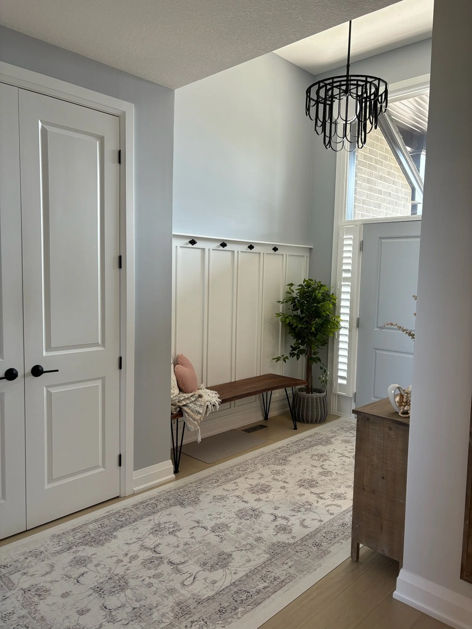 Interior view of a modern home entryway with a white door, hanging black chandelier, window with shutters, built-in white wall panel, and a bench with pillows. There is a decorative rug on the wooden floor and a potted plant next to the window.