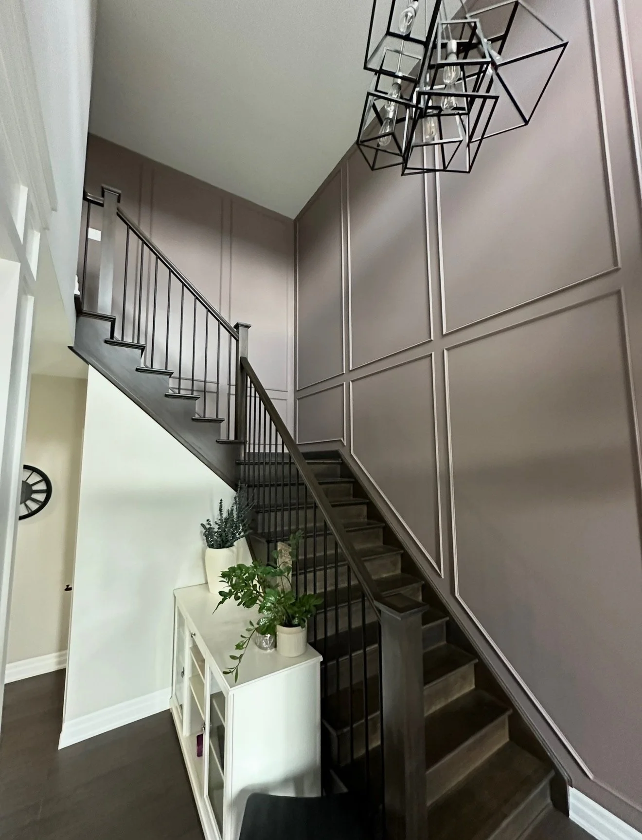 Interior view of a staircase with black railing and dark wooden steps, wainscoting on the wall, modern black geometric chandelier hanging, white console table with potted plants beneath the staircase.