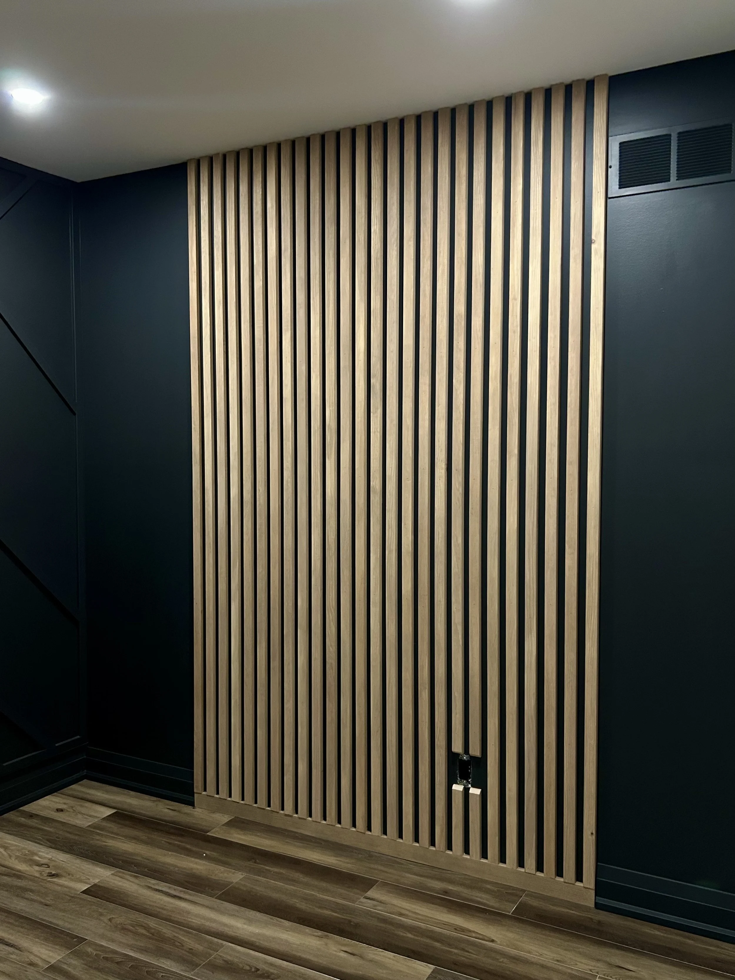 Interior of a room featuring a wall with vertical wooden slats and dark-colored walls on either side, with a section of wooden floor visible.