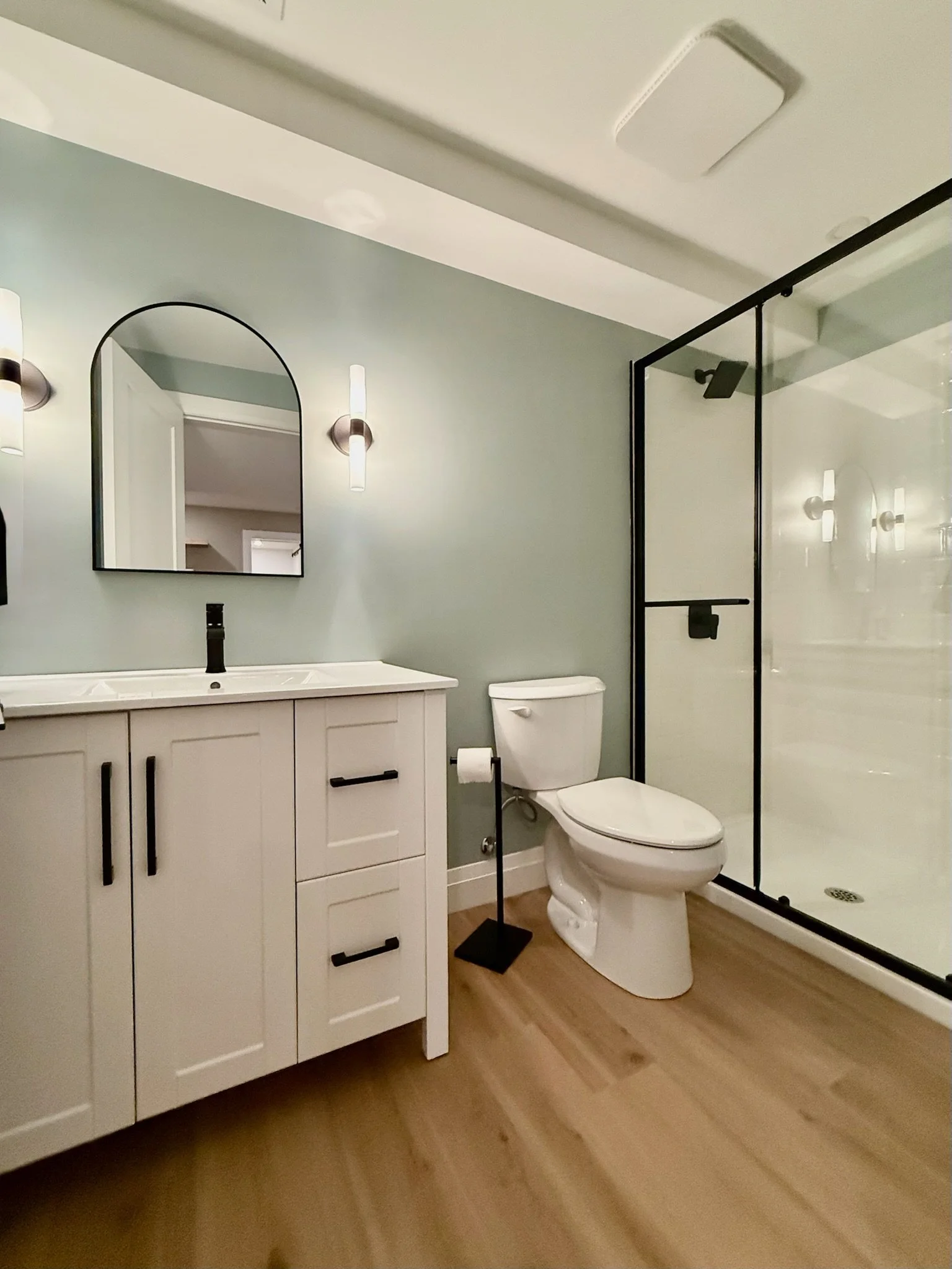 A modern bathroom with a white vanity, black handles, and a rectangular mirror above. There are wall-mounted lights on either side of the mirror. there is a glass shower enclosure with a black frame. The flooring is LVP