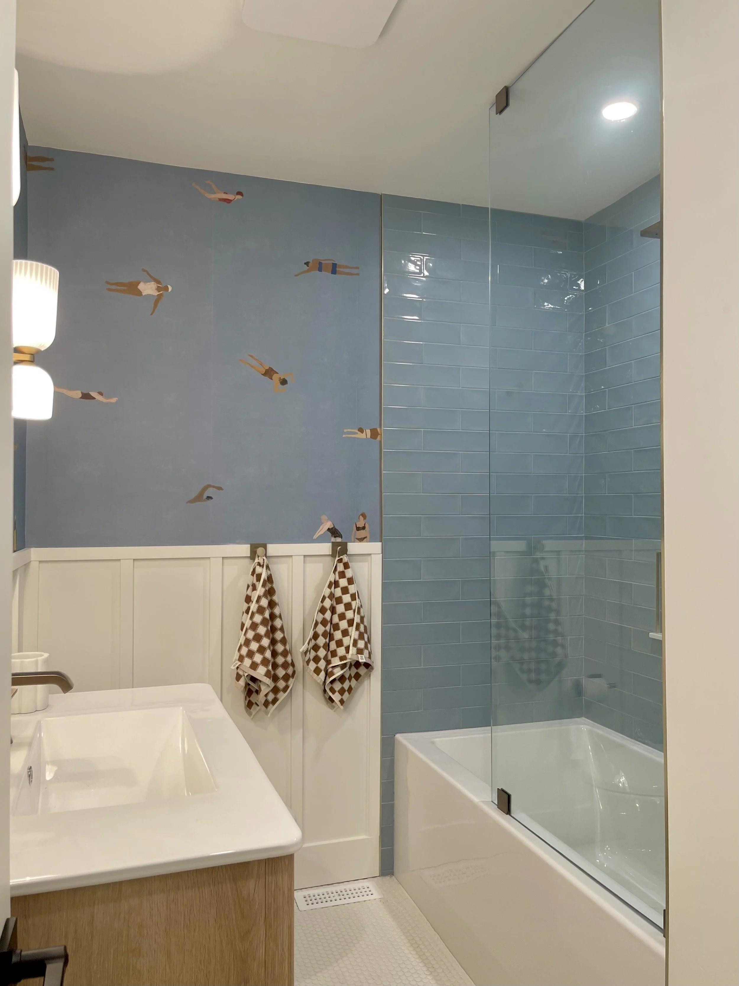 Modern bathroom with a white sink and wooden vanity, a blue tiled shower with a glass door, and a light blue wall with illustrated people swimming.