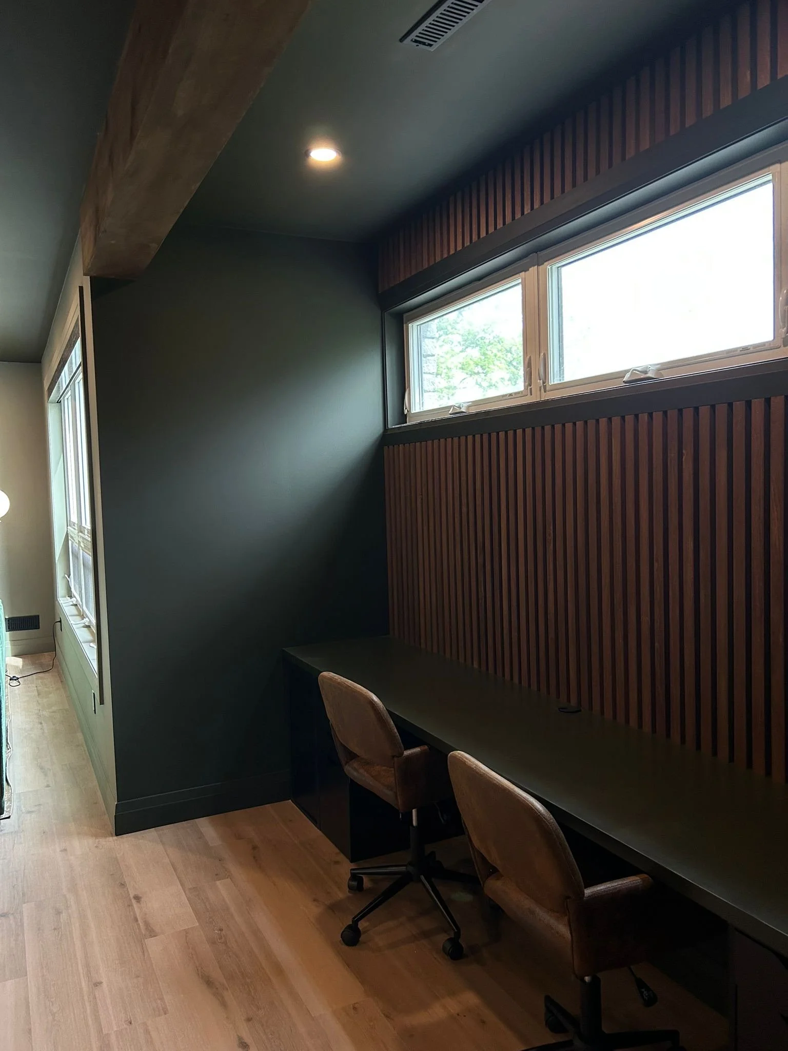 Interior of a modern basement gaming zone and office with a long black desk, three brown chairs, dark green walls, wood paneling, and windows letting in natural light.
