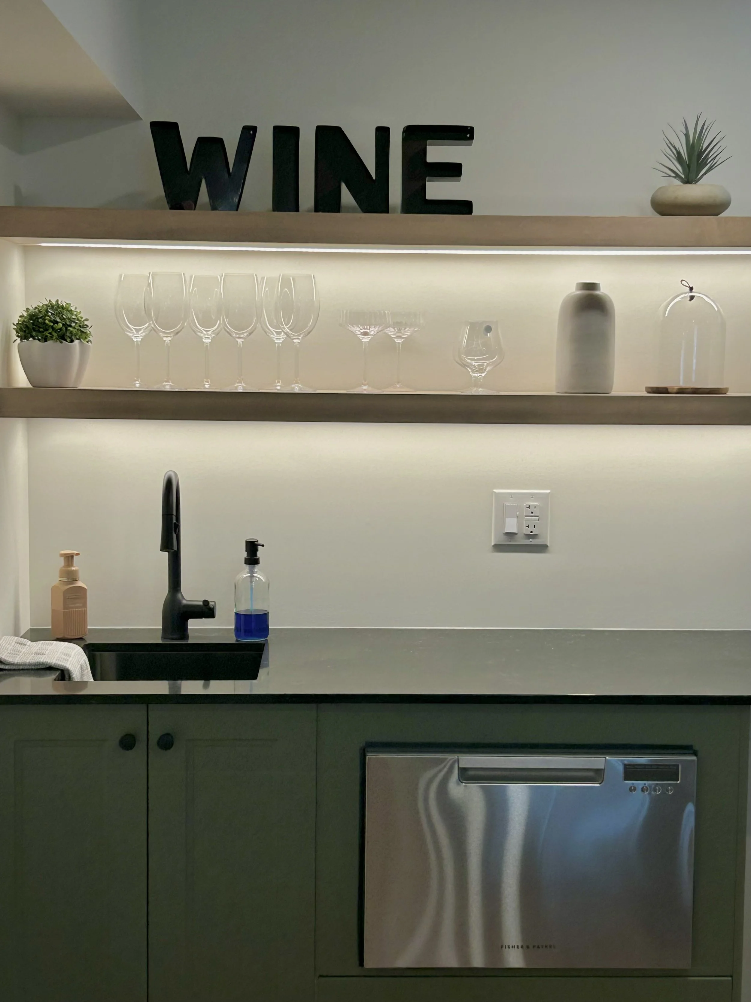 Modern kitchen with green cabinets, black faucet, soap dispenser, wine glasses, decorative vases, and a shelf with "WINE" letters and a potted plant.