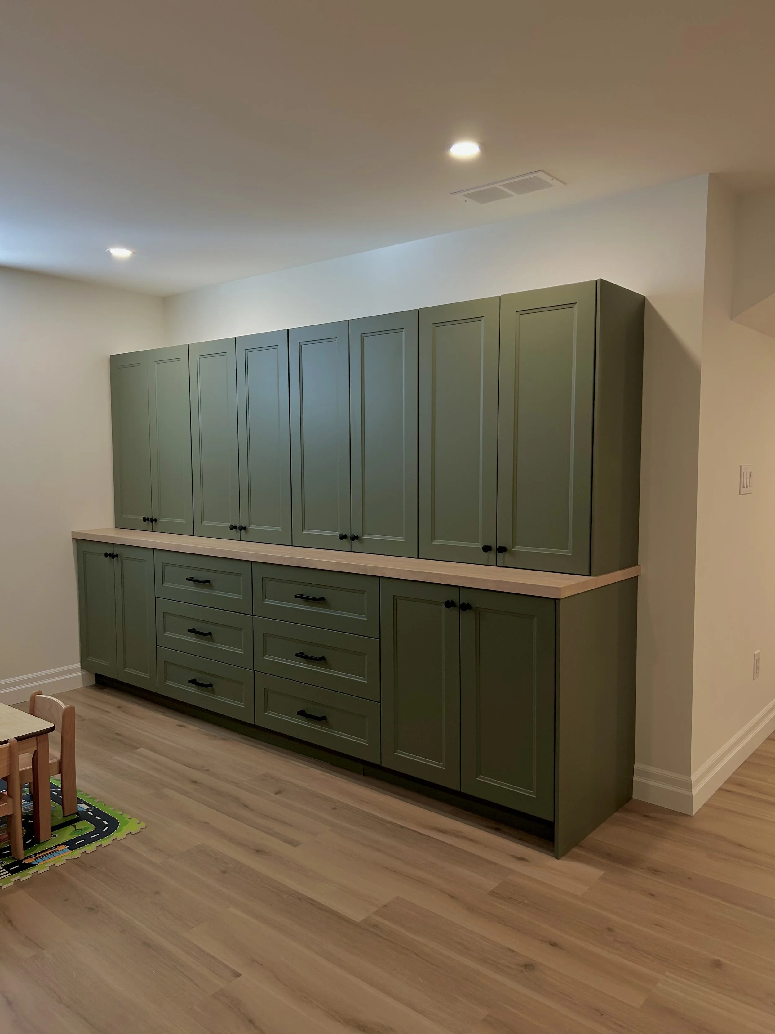 Green ikea cabinets with black handles for a built in for kid storage, with a LVP floor, wooden counter and beige walls.