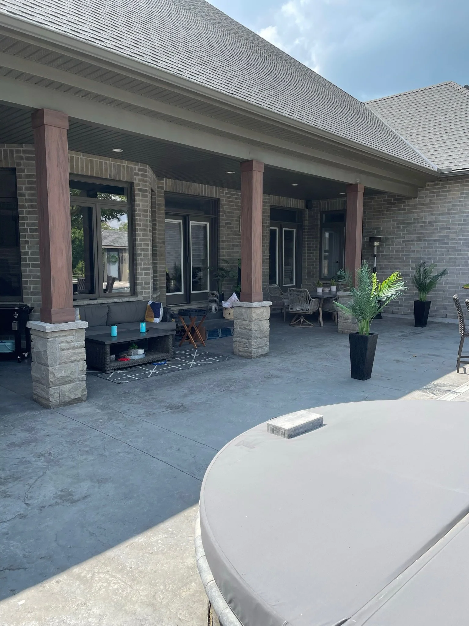 A backyard patio area with a brick house, large windows, several potted plants, outdoor furniture, and a covered porch with wooden posts and recessed lighting, under a partly cloudy sky.