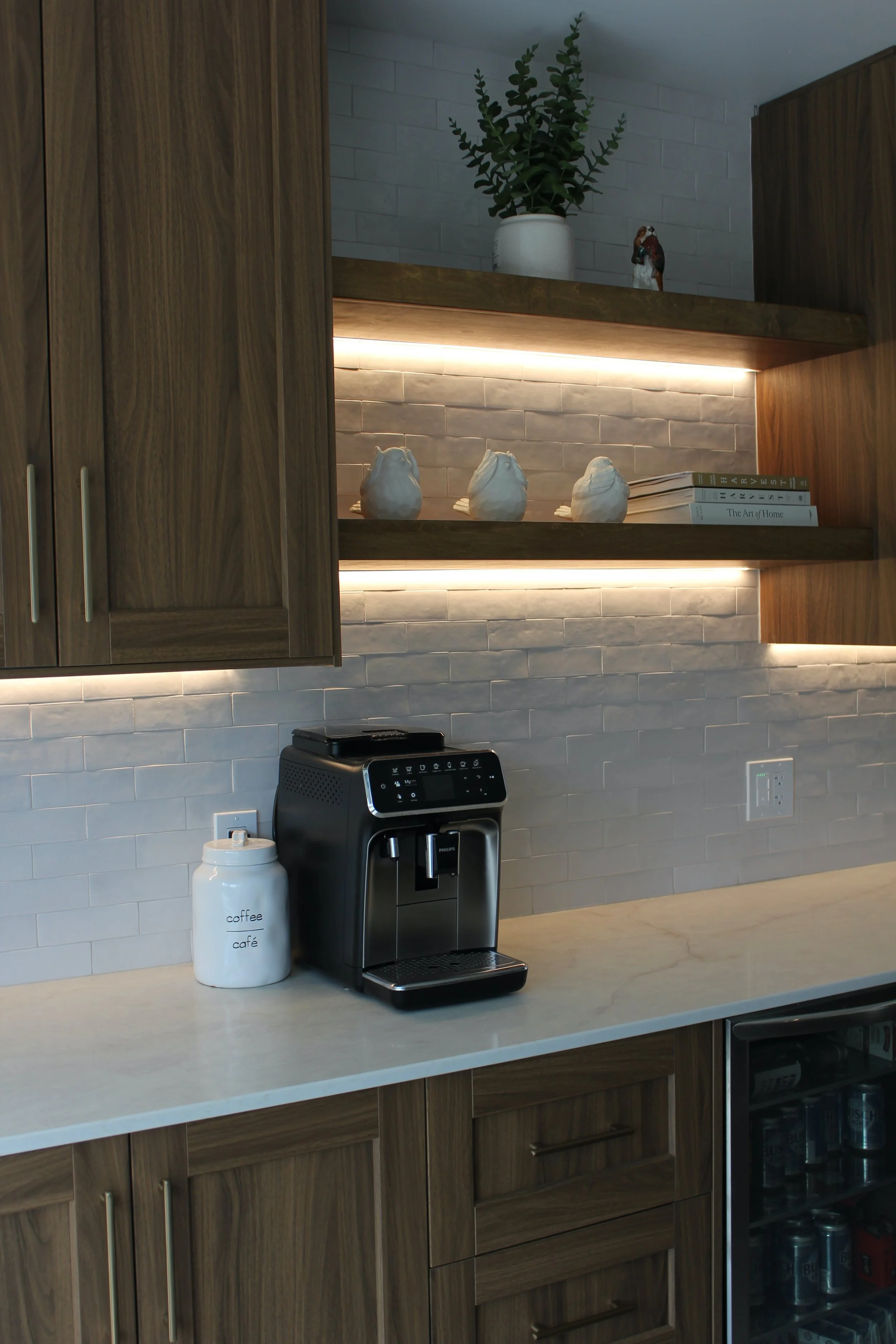 Kitchen countertop with a black coffee maker, a white coffee canister labeled 'coffee café,' a small white ceramic container, wooden cabinets, and open shelves with decorative items and books. There's a white tiled backsplash with LED lighting undern