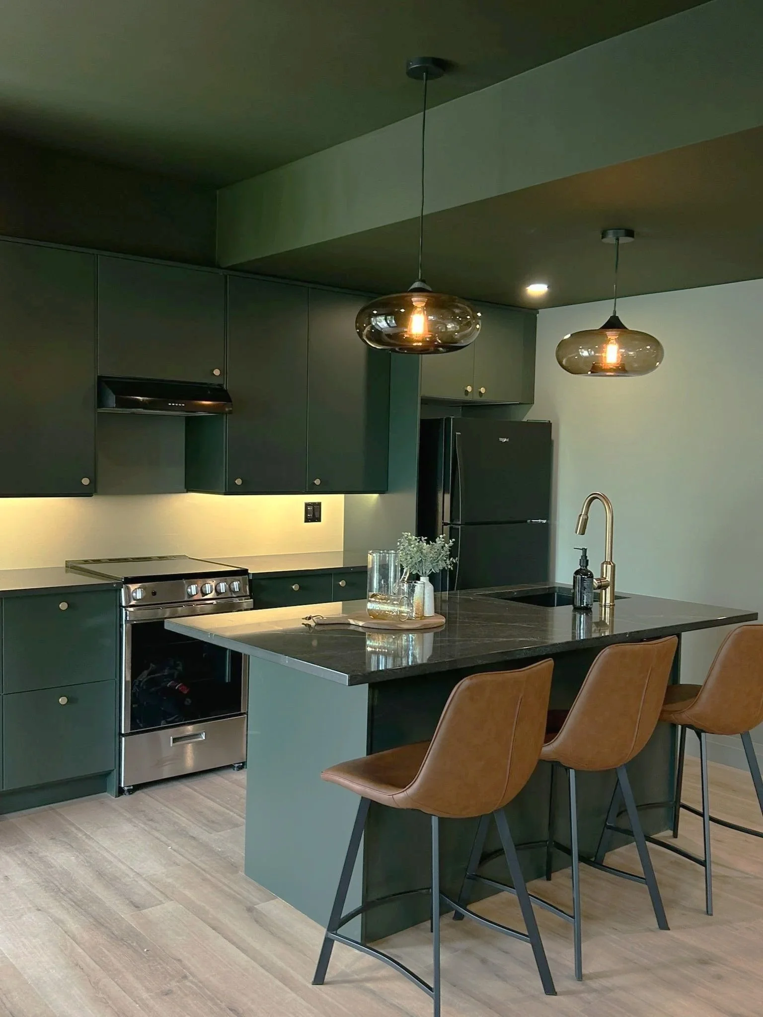 Modern Ikea Cabinet kitchen with dark green cabinets, black appliances, a black Hanstone Markina Quartz with wooden chairs, pendant lights, and a LVP floor.