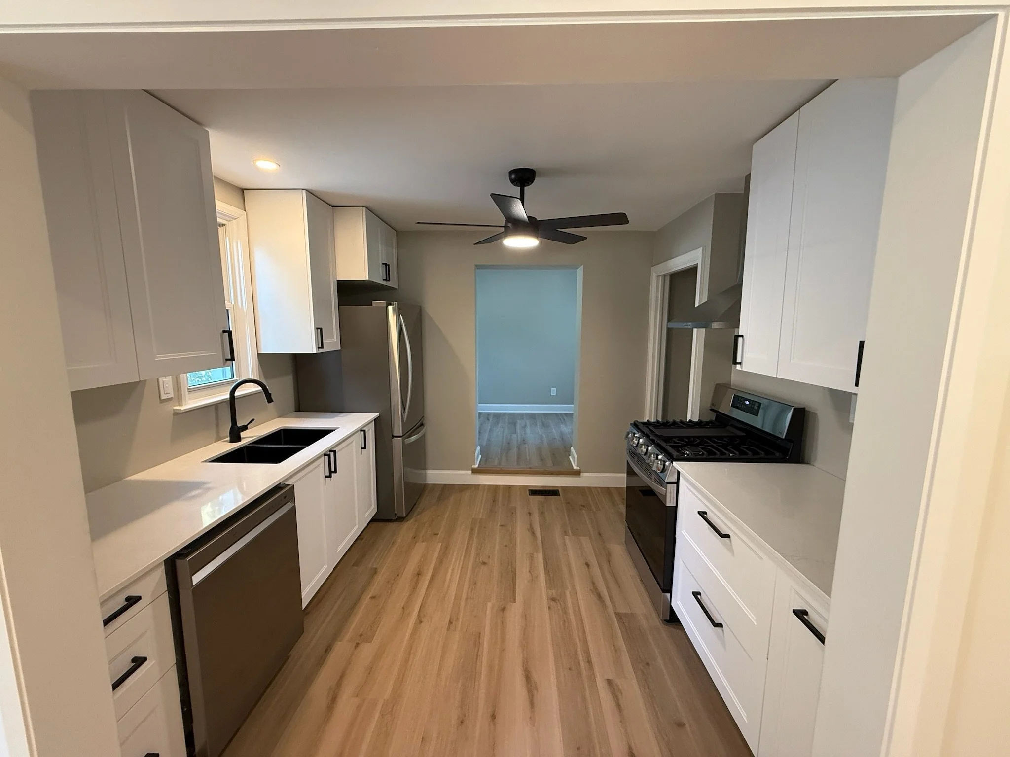 A modern kitchen with white cabinets, black hardware, a black range, stainless steel refrigerator, and a black ceiling fan with a light. Wooden flooring and a small window above the sink.
