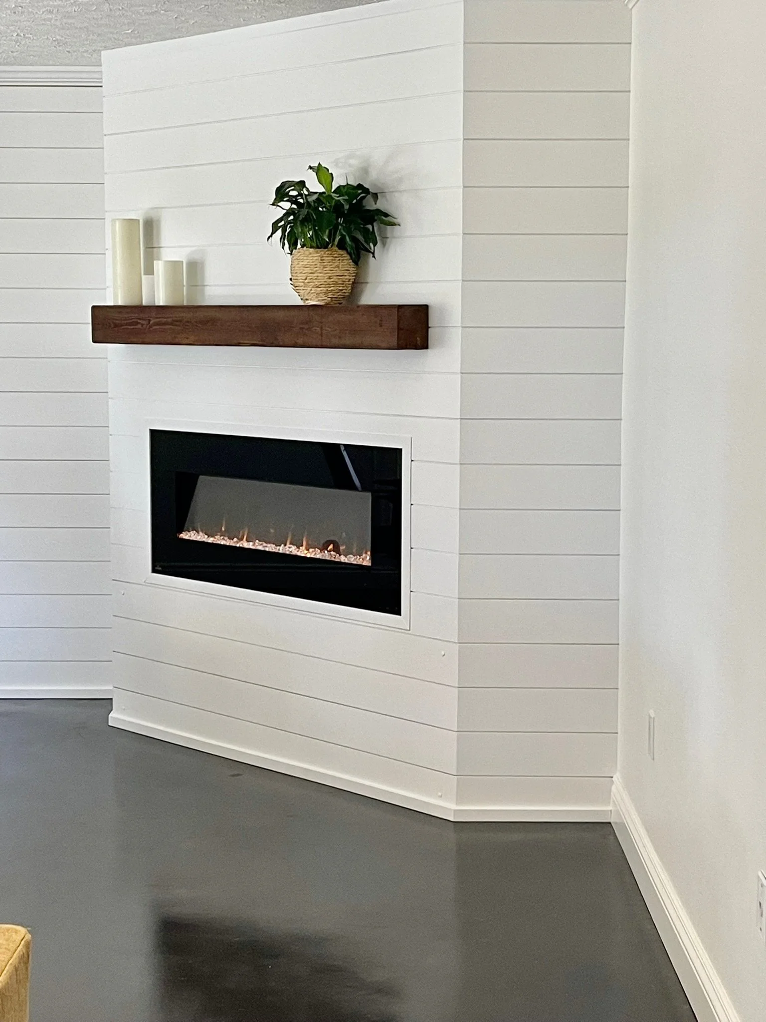 A living room corner with white shiplap walls, a fireplace, and a dark gray floor. A wooden mantel holds a potted plant and three candles. Part of a beige sofa is visible in the bottom left corner.