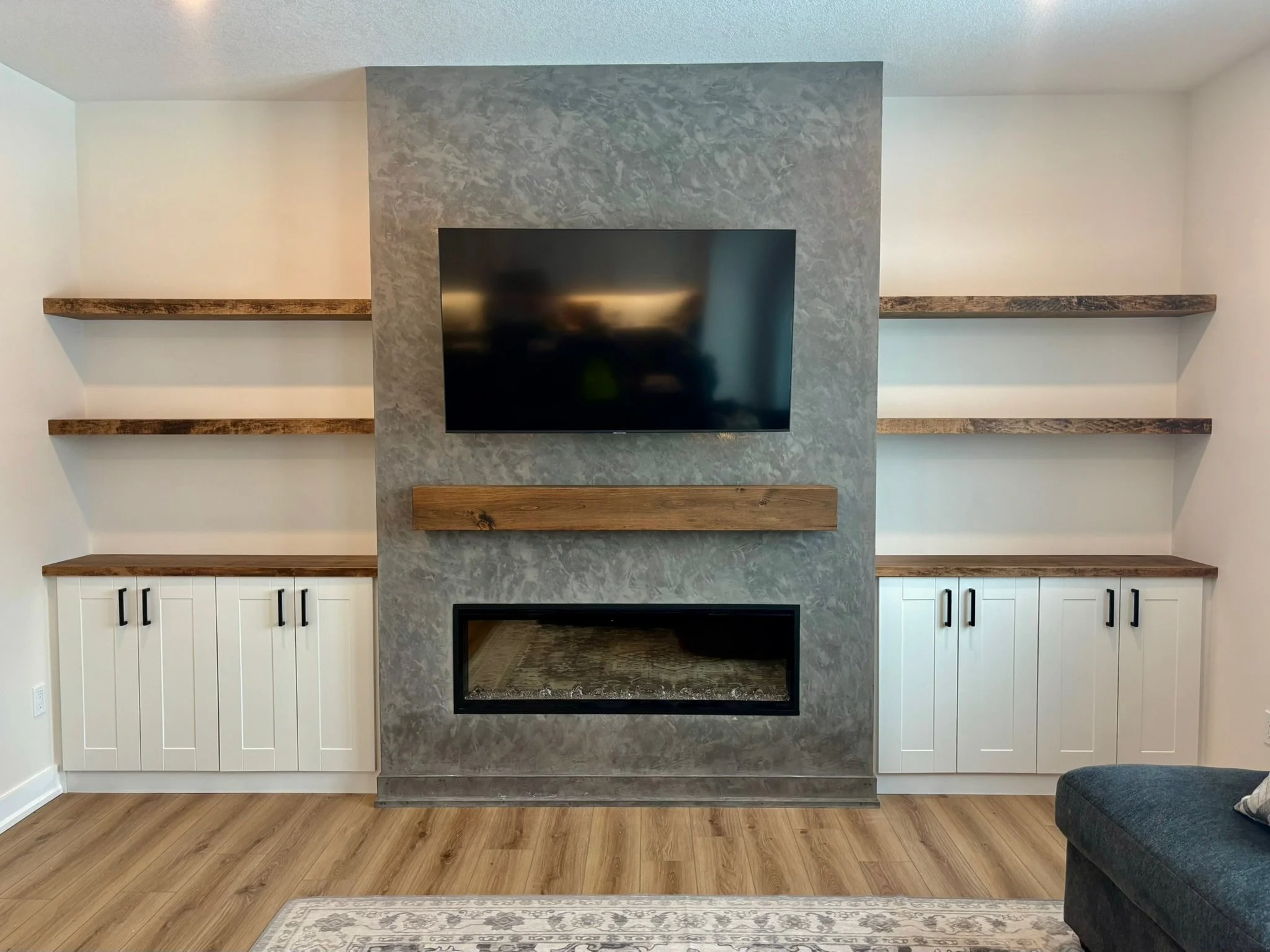 Living room with a gray textured wall and a flat-screen TV mounted on it, flanked by wooden shelves, white cabinets below, and hardwood floors.