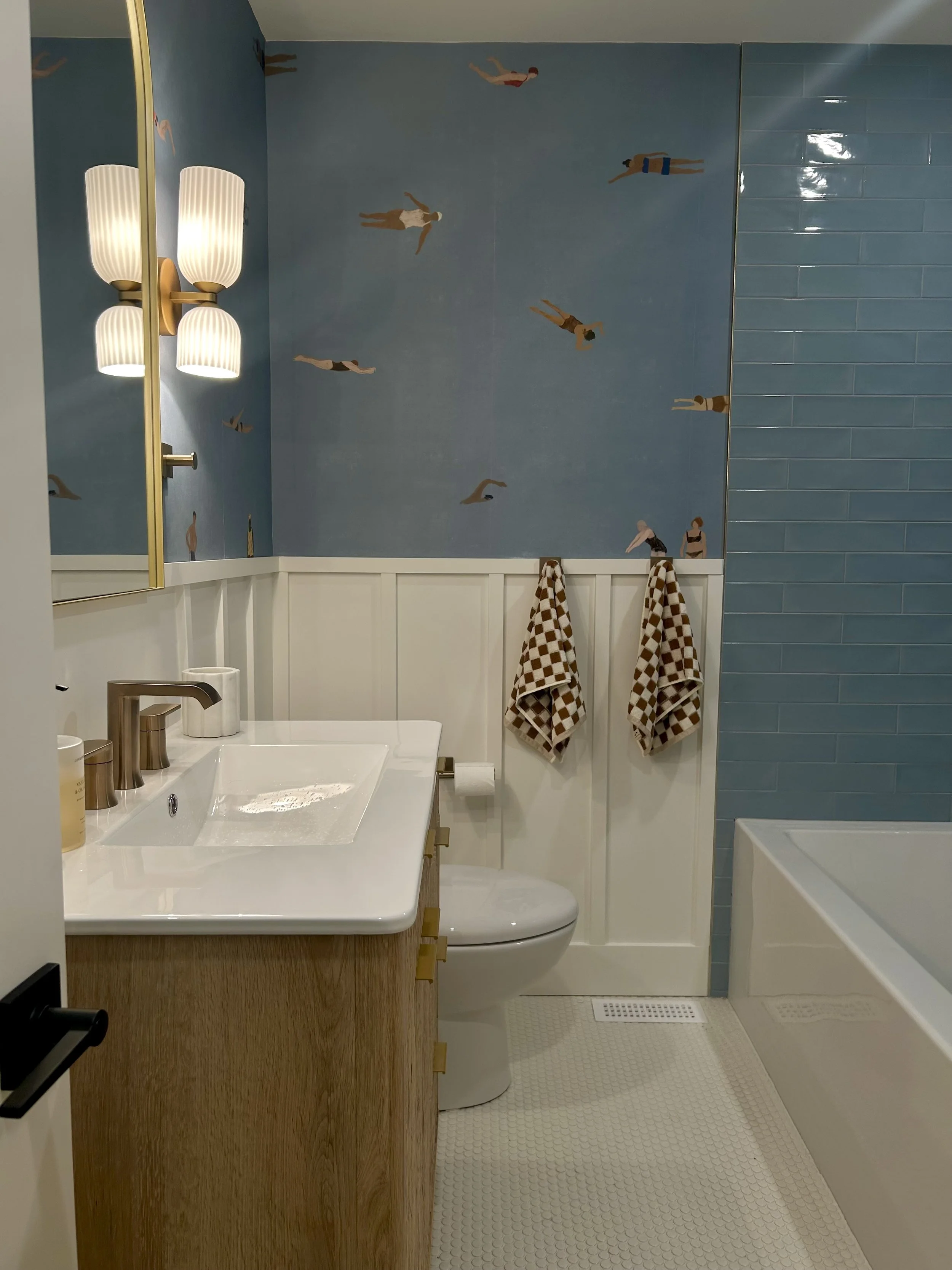 A bathroom with a blue wallpaper of  painted figures of people swimming, a white vanity with a sink and a toilet, checkered brown and beige towels hanging, a wall-mounted mirror with a light fixture, and blue subway tile on part of the wall.