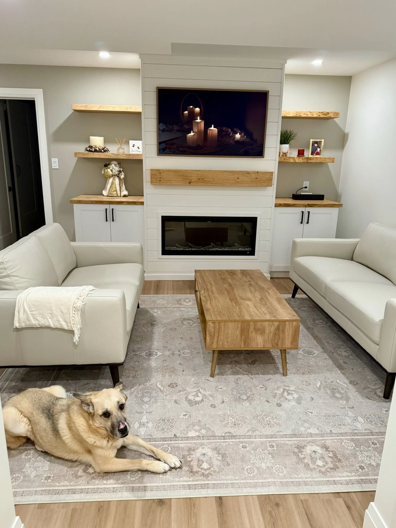 Living room with two white couches, a wooden coffee table, a fireplace, wall-mounted TV, built in cabinets and wooden shelves with decor items. A dog is lying on a patterned rug on the floor.