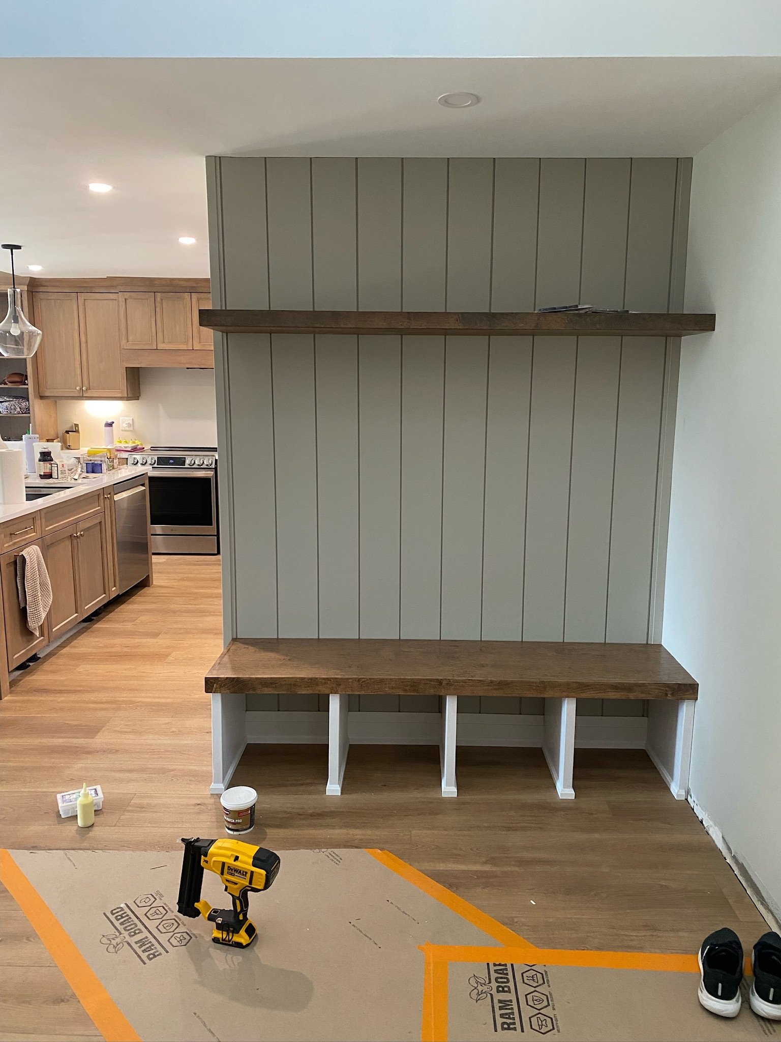 A room under renovation with a partially installed wooden and beadboard wall. Construction tools and supplies, including a cordless drill, paint, and shoes, are on the floor. The kitchen area with wooden cabinets and appliances is visible in the back