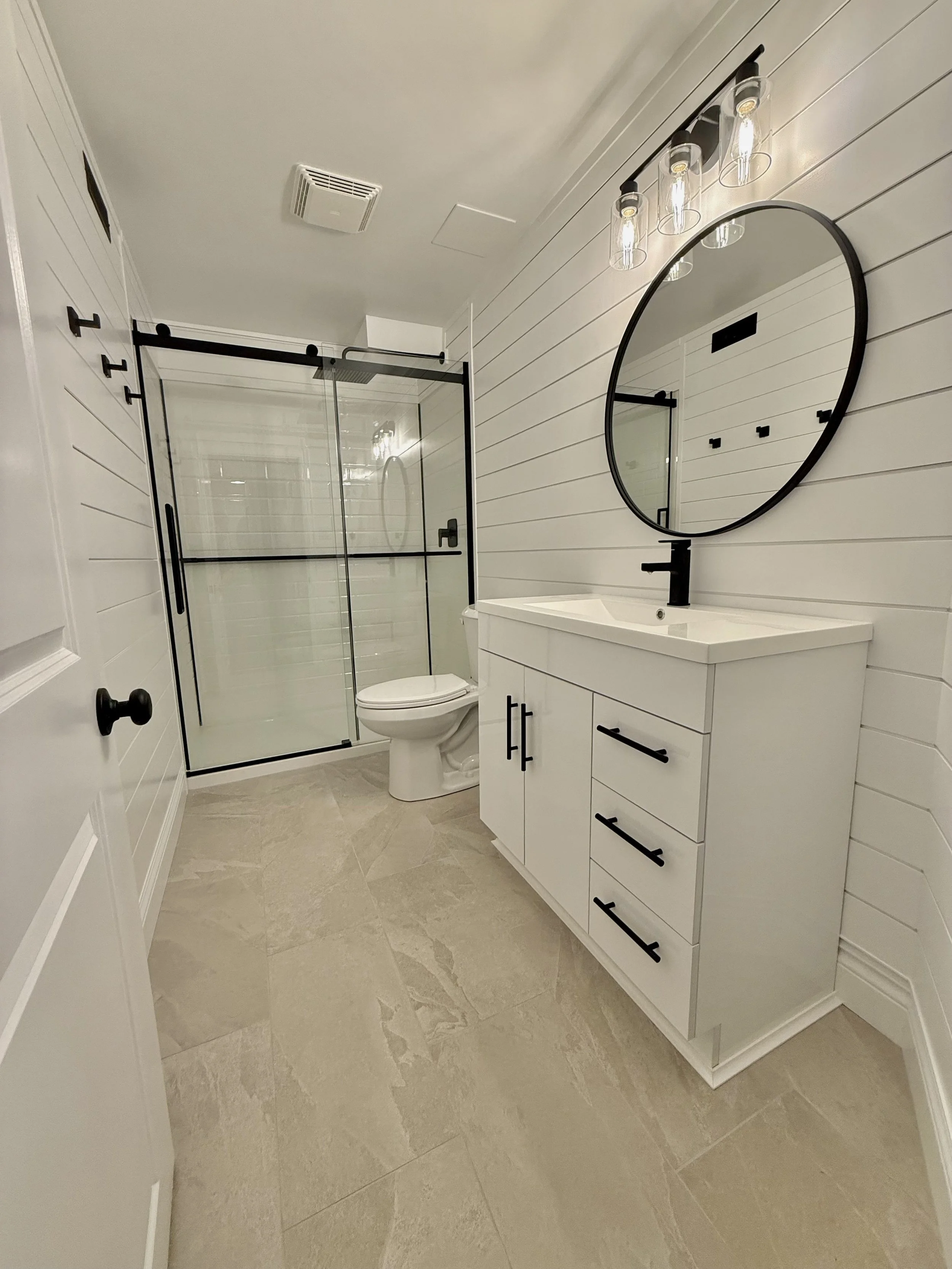 A modern bathroom with a glass shower enclosure, white vanity with black hardware, round mirror, black light fixture, toilet, and beige tiled floor and walls.