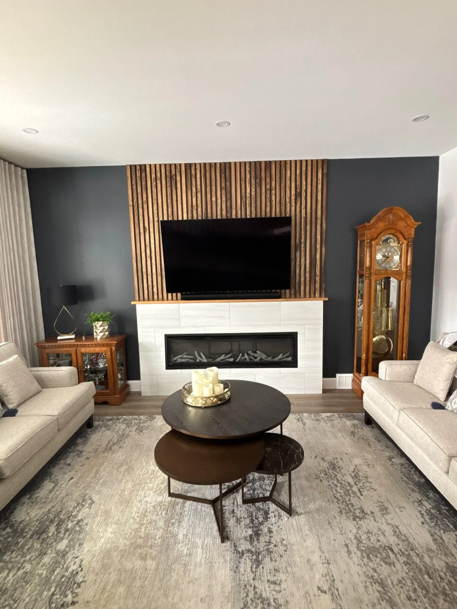 Living room a wall-mounted TV on a wood slate wall above a fireplace with white stone surround. The wall behind the TV is dark gray, and there's a large beige area rug on the floor and 2 beige sofas.
