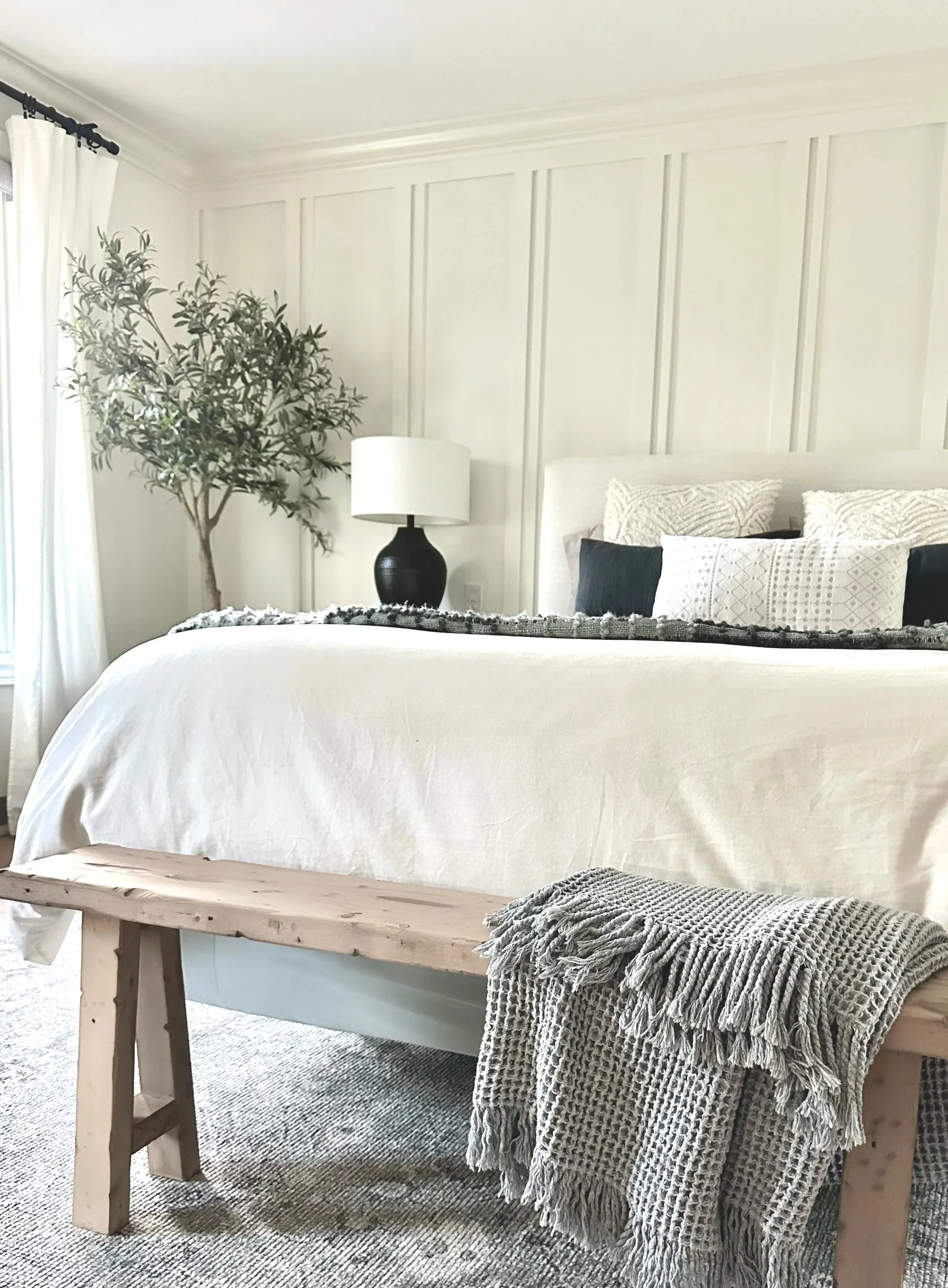 A bedroom with a bed, decorated in neutral tones. The bed has white, patterned, and dark pillows. A textured blanket is draped over a wooden bench at the foot of the bed. To the left, there is a large green plant and a black table lamp with a white shade. The wall features panel molding, and there are white curtains by a window.